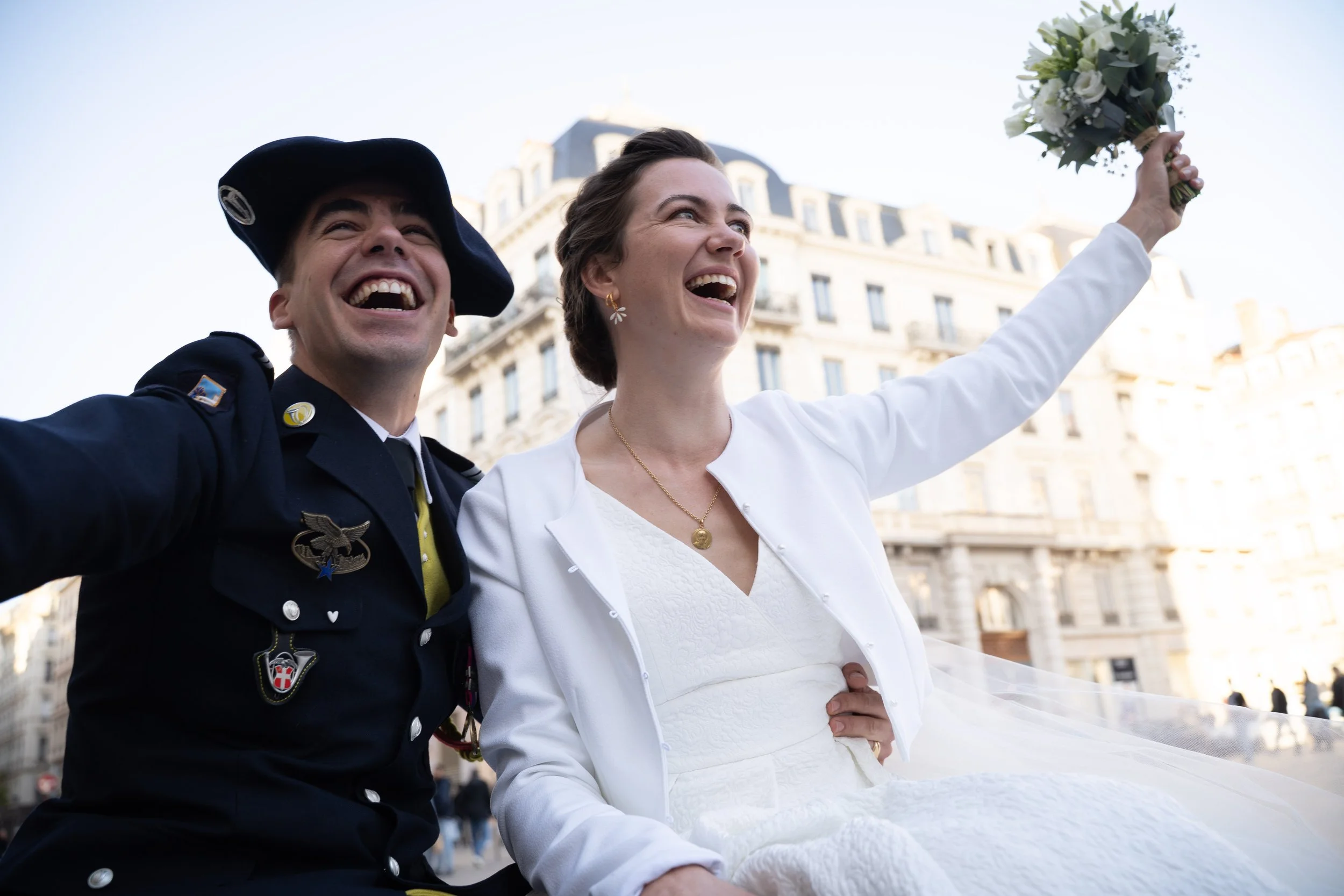 Un homme en uniforme militaire avec un chapeau noir et une femme en robe blanche souriant ensemble, la femme tient un bouquet de fleurs en l'air dans une ville en arrière-plan, probablement lors d'une célébration ou d'un mariage.