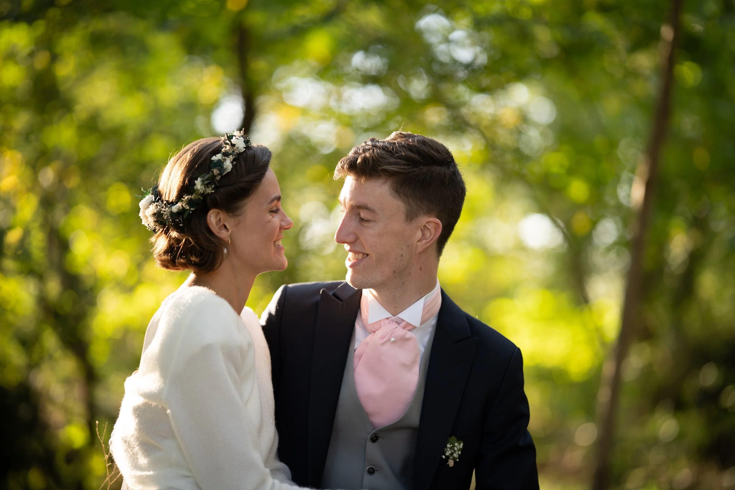 Un couple en tenue de mariage, la femme portant une couronne de fleurs, fait face l'un à l'autre sous un arbre en pleine nature, souriant et se regardant tendrement.