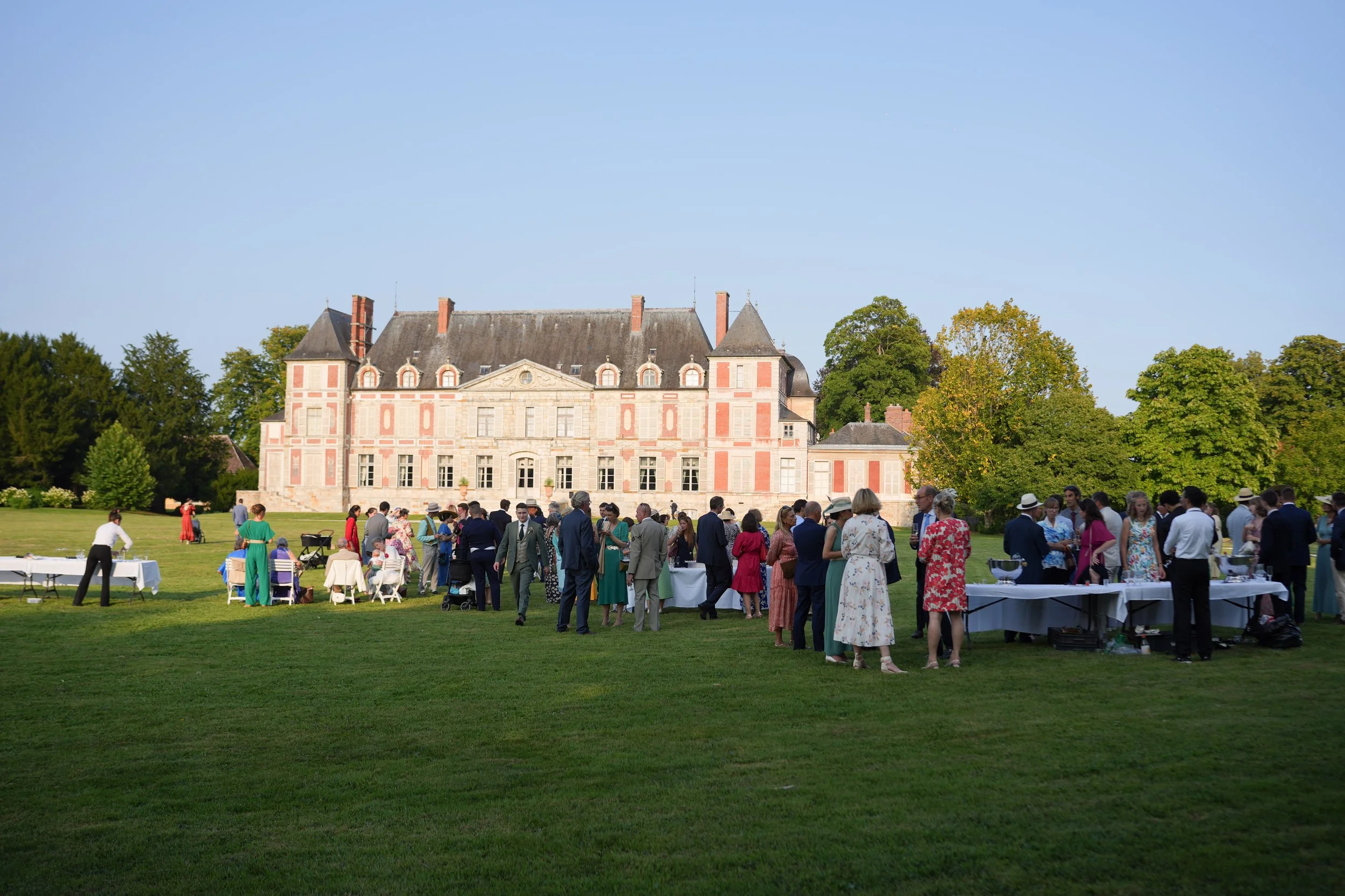 Un groupe de personnes habillées élégamment assistent à un événement en plein air devant un grand château. La scène se déroule en été, avec des arbres verts au fond.