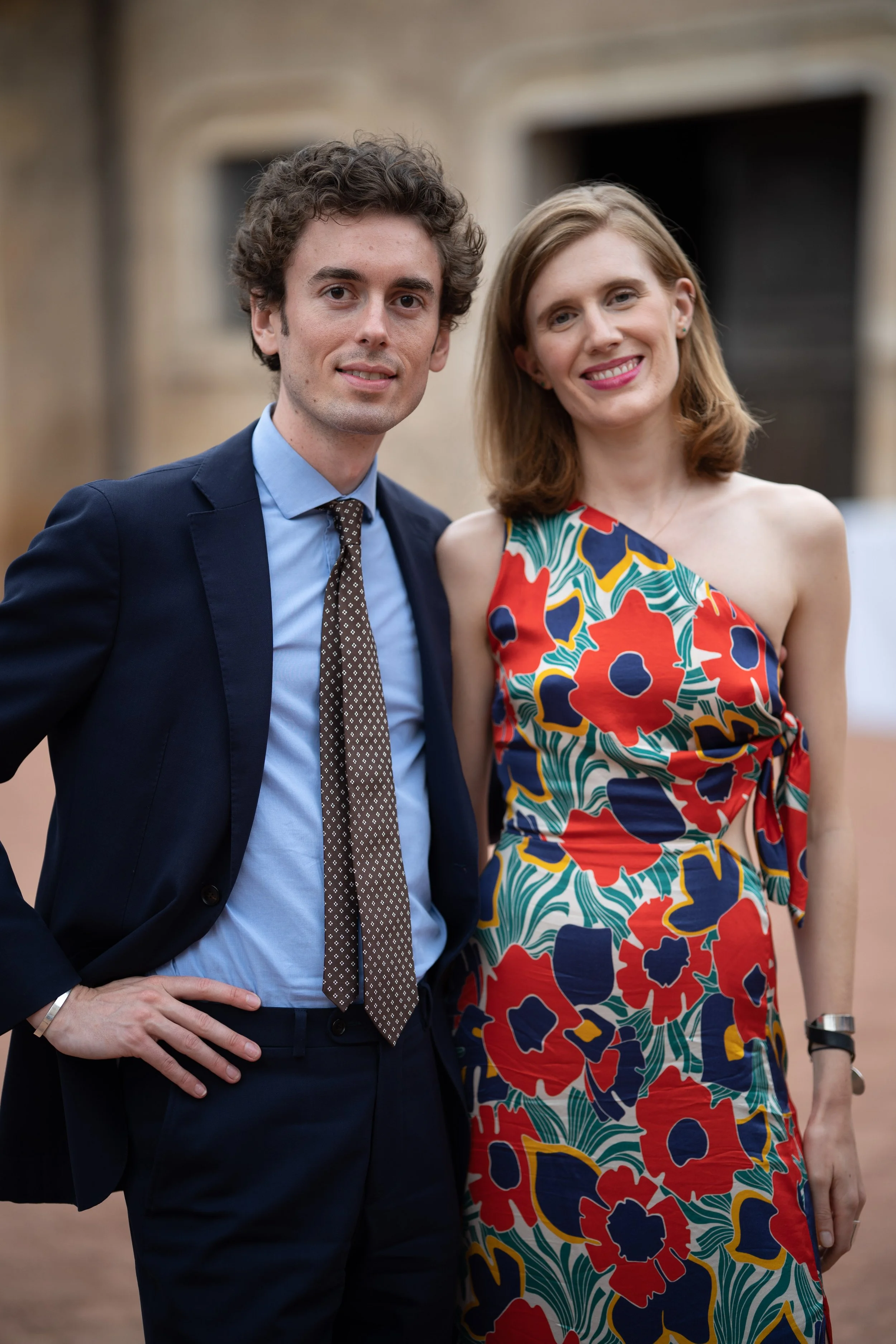 Un homme en costume sombre et une femme en robe colorée posent ensemble à l'extérieur, sourire et regardant l'appareil photo.