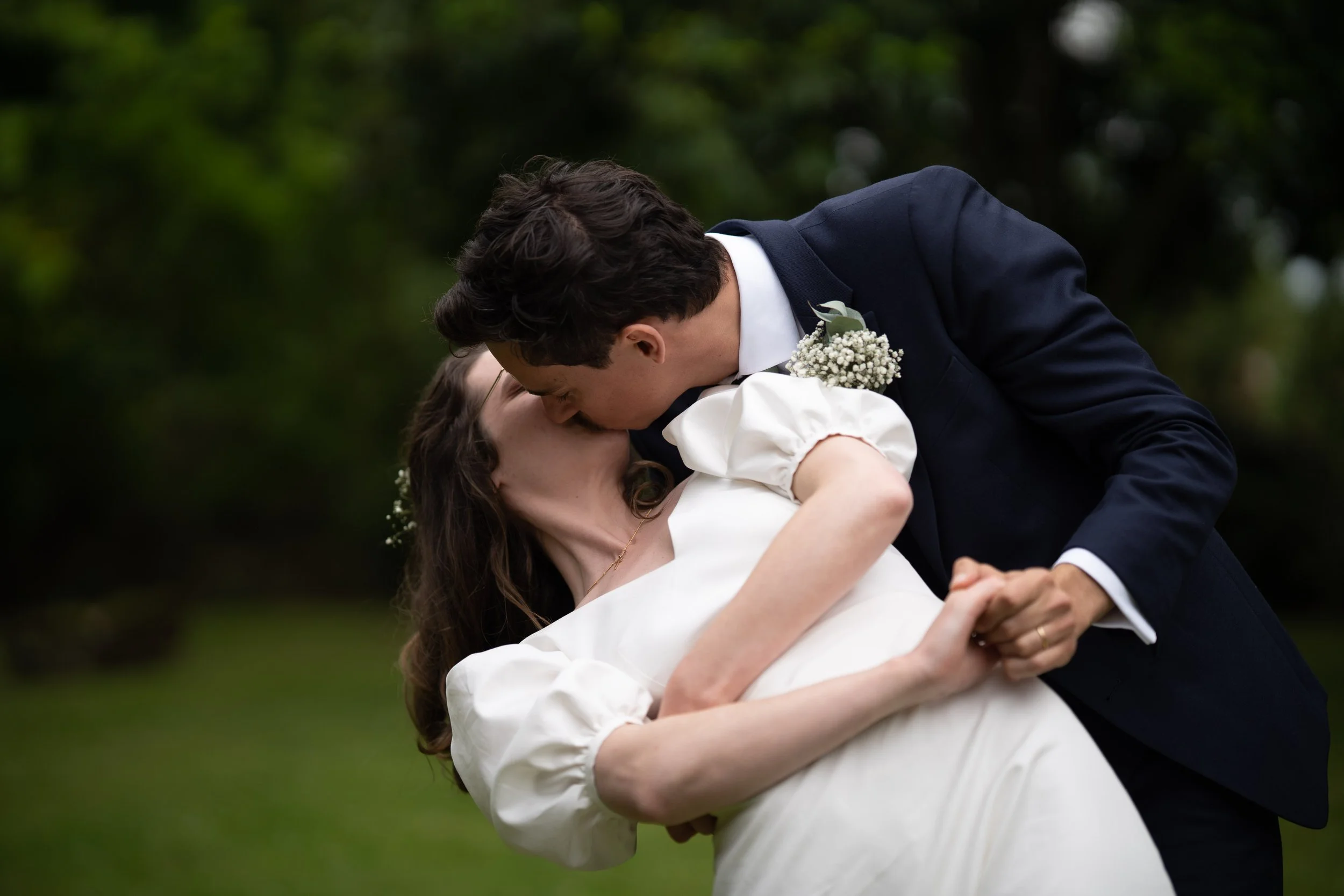 Un couple en costume de mariage danse en plein air, la femme porte une robe blanche et des fleurs dans les cheveux, l'homme porte un costume sombre avec une boutonnière, derrière un fond de nature avec des arbres.