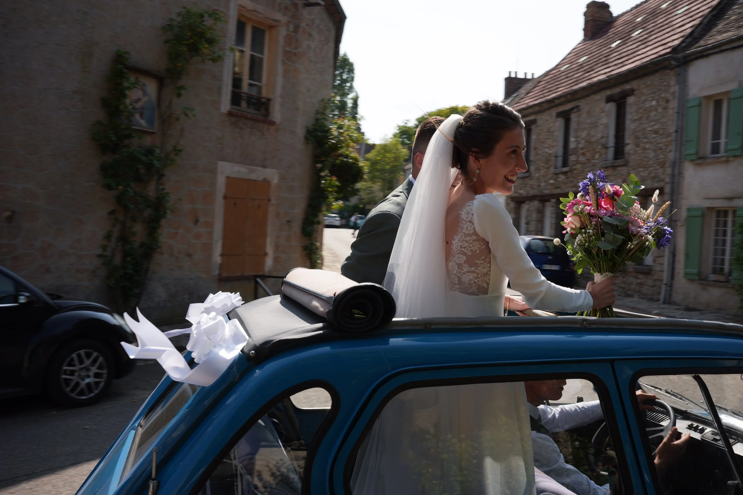 Une femme en robe de mariage souriante, portant un voile, tenant un bouquet de fleurs, à l'arrière d'une voiture ancienne décorée de rubans blancs, dans une rue pavée avec des bâtiments en pierre.