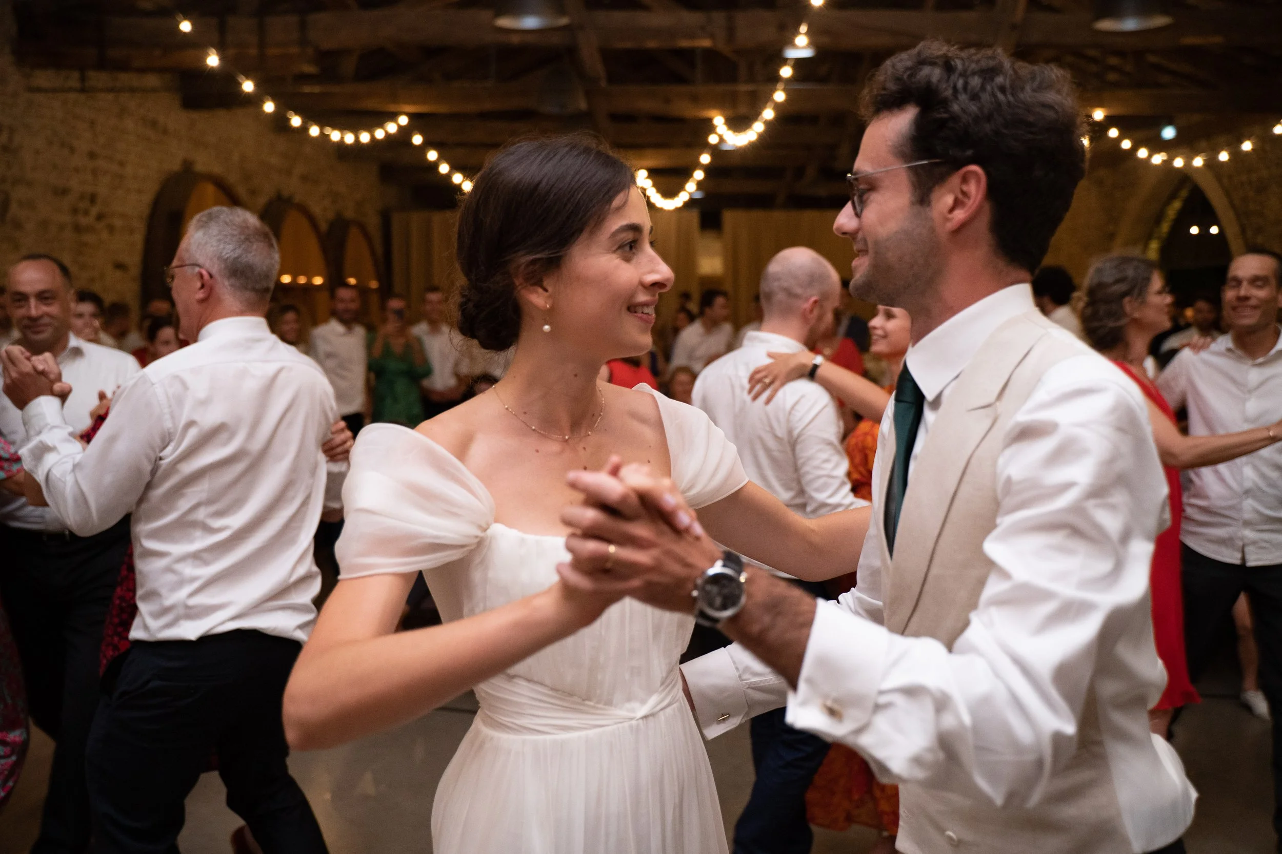 Un couple danse lors d'une fête dans une salle avec des lumières suspendues et un mur en briques, entouré d'autres personnes qui dansent ou regardent.