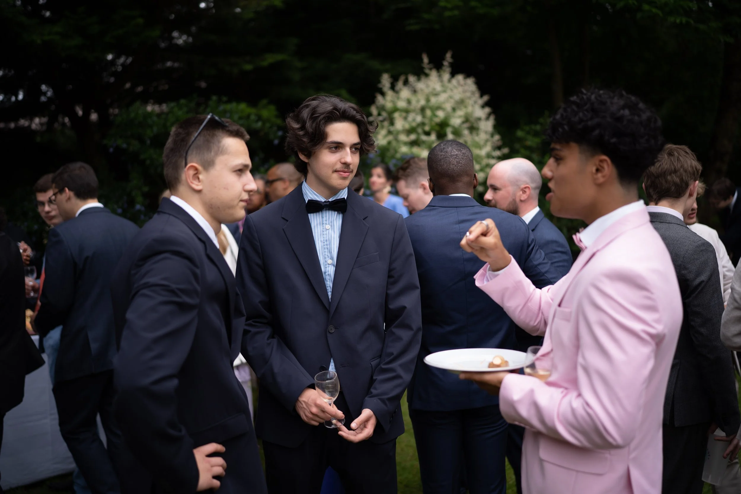 Groupe de jeunes hommes en costume lors d'une fête en plein air, discutant et se tenant par la main.