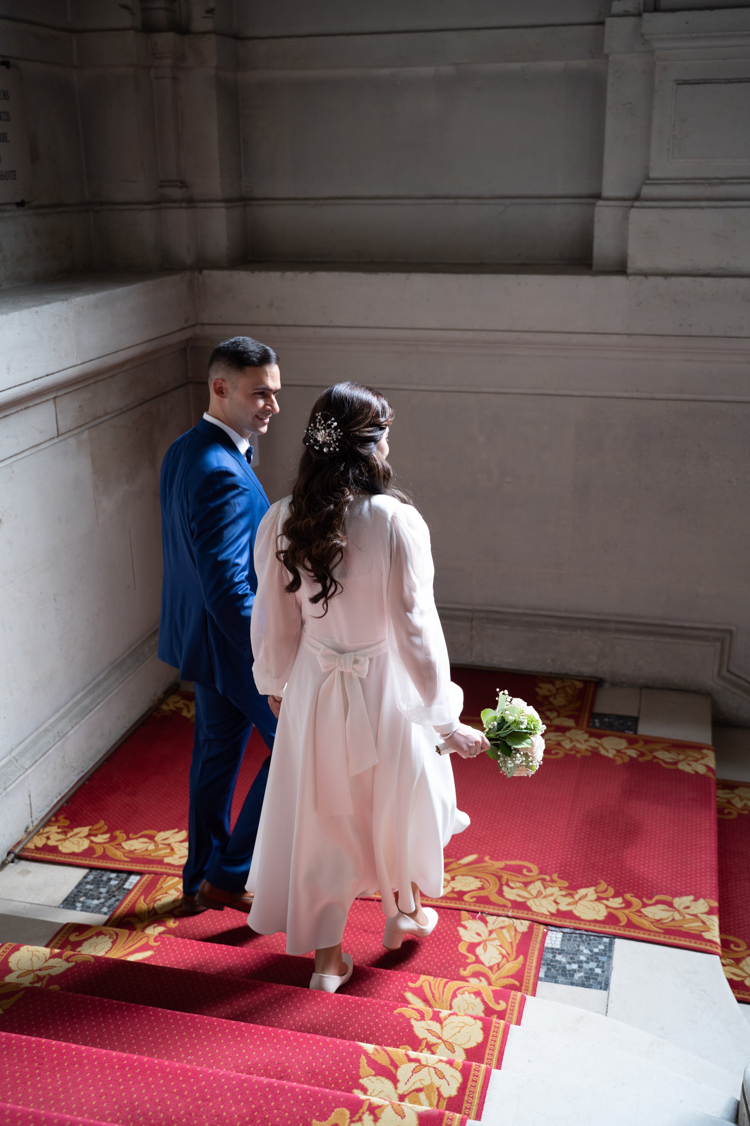 Un couple habillé en mariage, la femme tenant un bouquet de fleurs, descend les escaliers dans un lieu historique ou religieux en pierre, l'ambiance est festive et élégante.