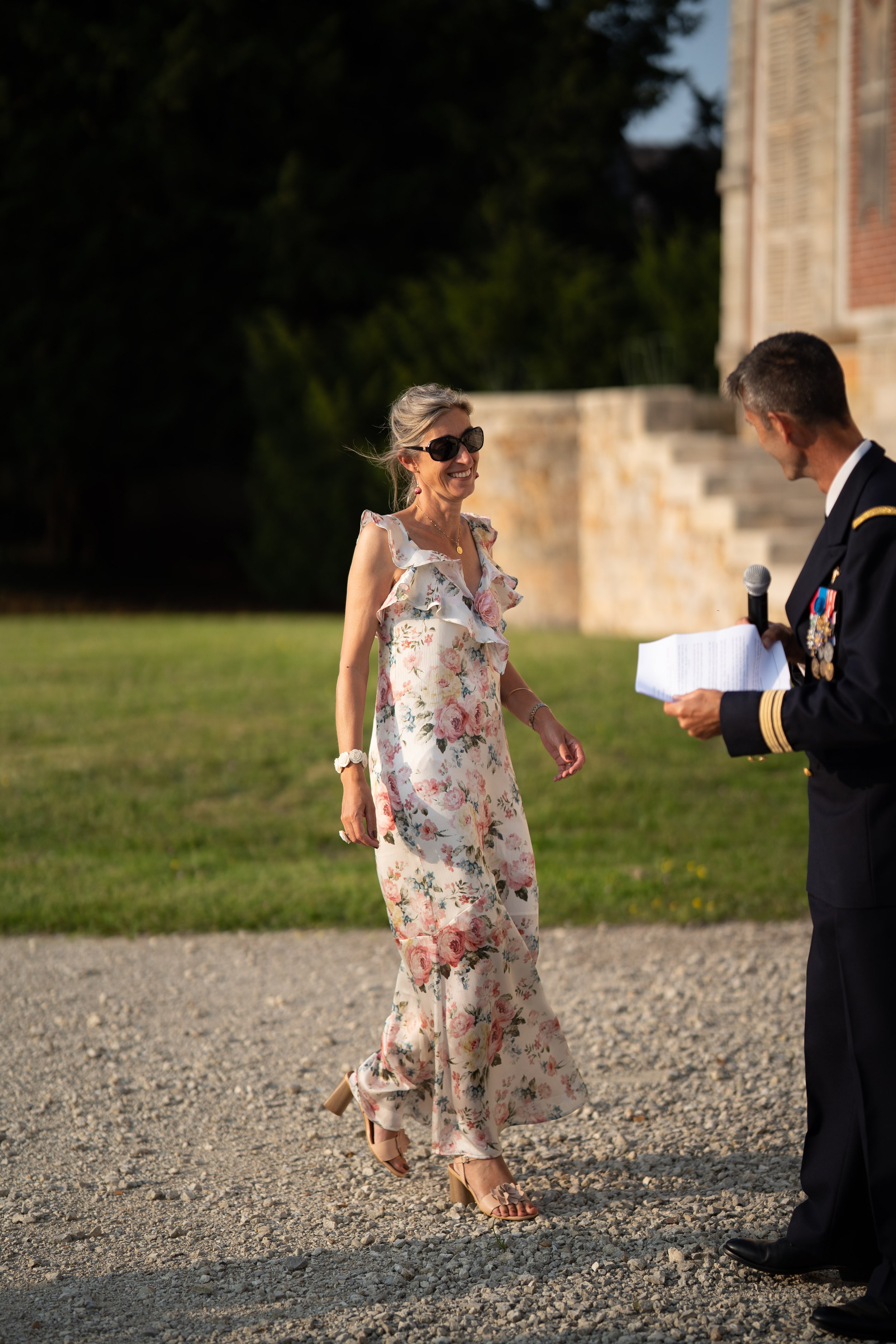 Une femme en robe longue florale sourit en regardant un homme en uniforme qui lui remet un document, lors d'une cérémonie en plein air au coucher du soleil.