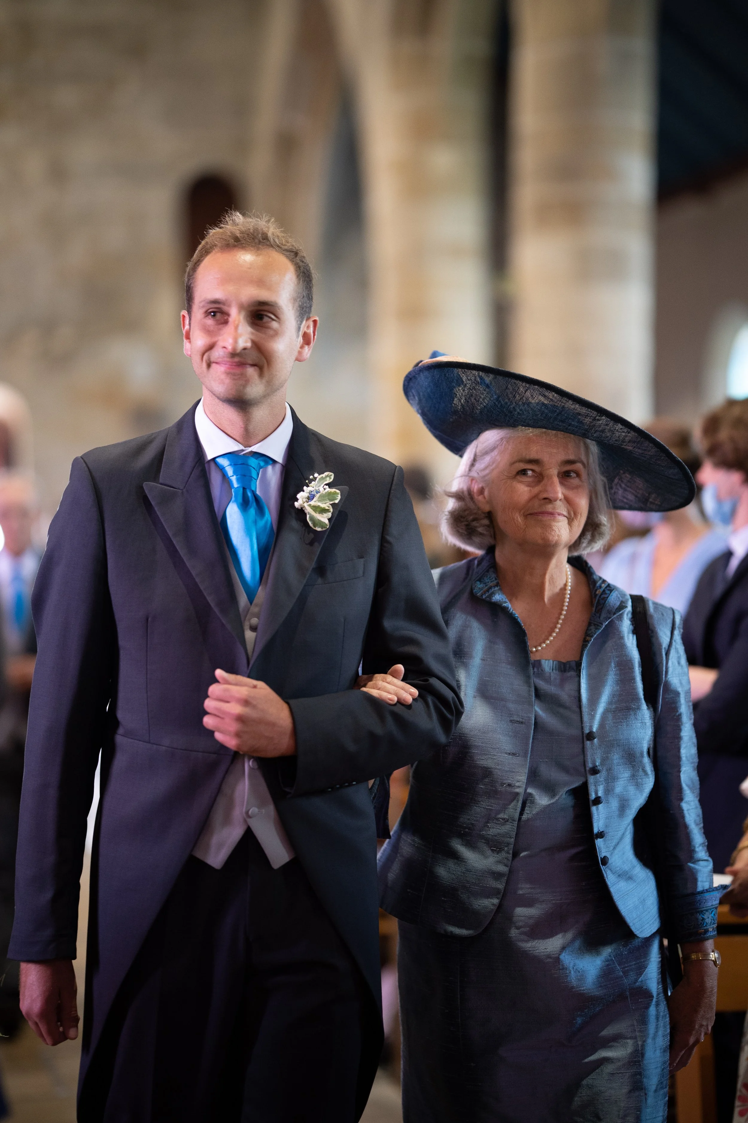 Un homme en costume de mariage et une femme âgée en robe élégante portant un large chapeau, se tenant par le bras lors d'une cérémonie dans une église.