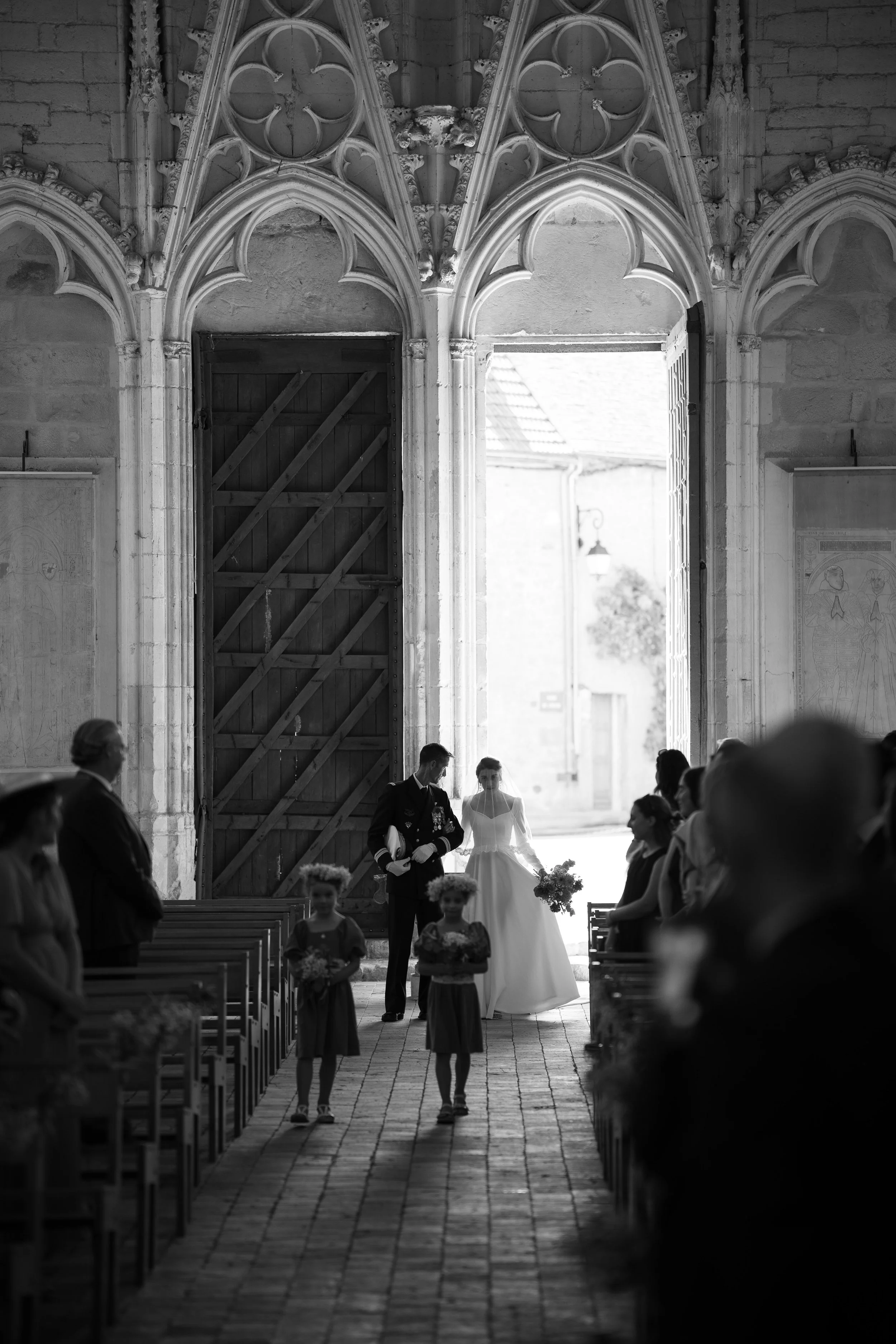 Une cérémonie de mariage dans une église avec des invités assis de part et d'autre, une mariée vêtue en blanc avec un bouquet, accompagnée d'un marié en uniforme, marchant dans l'allée centrale avant d'entrer dans la nef.