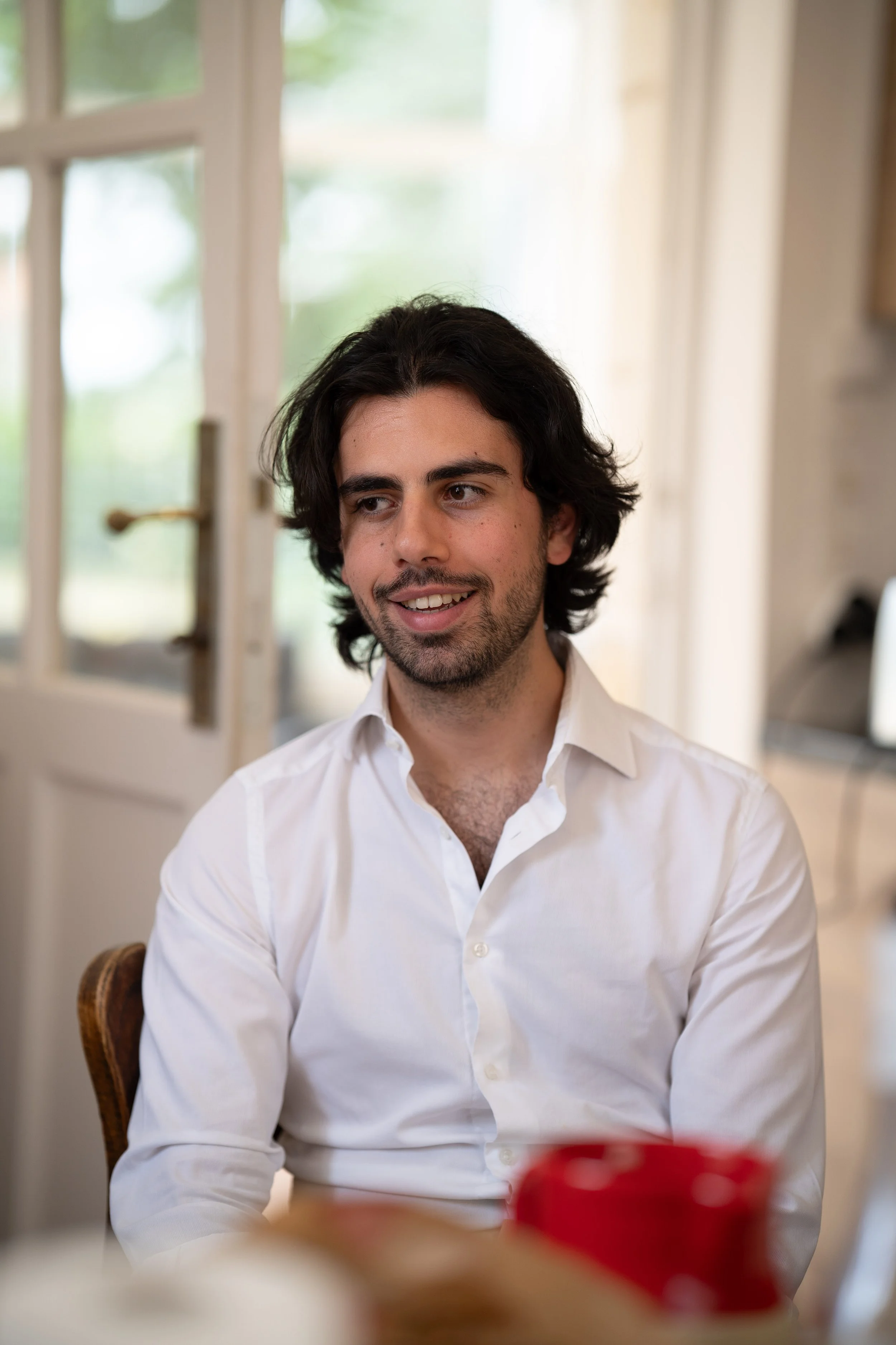 Jeune homme aux cheveux longs et barbe, souriant, portant une chemise blanche, assis dans une salle lumineuse avec une porte vitrée en arrière-plan.
