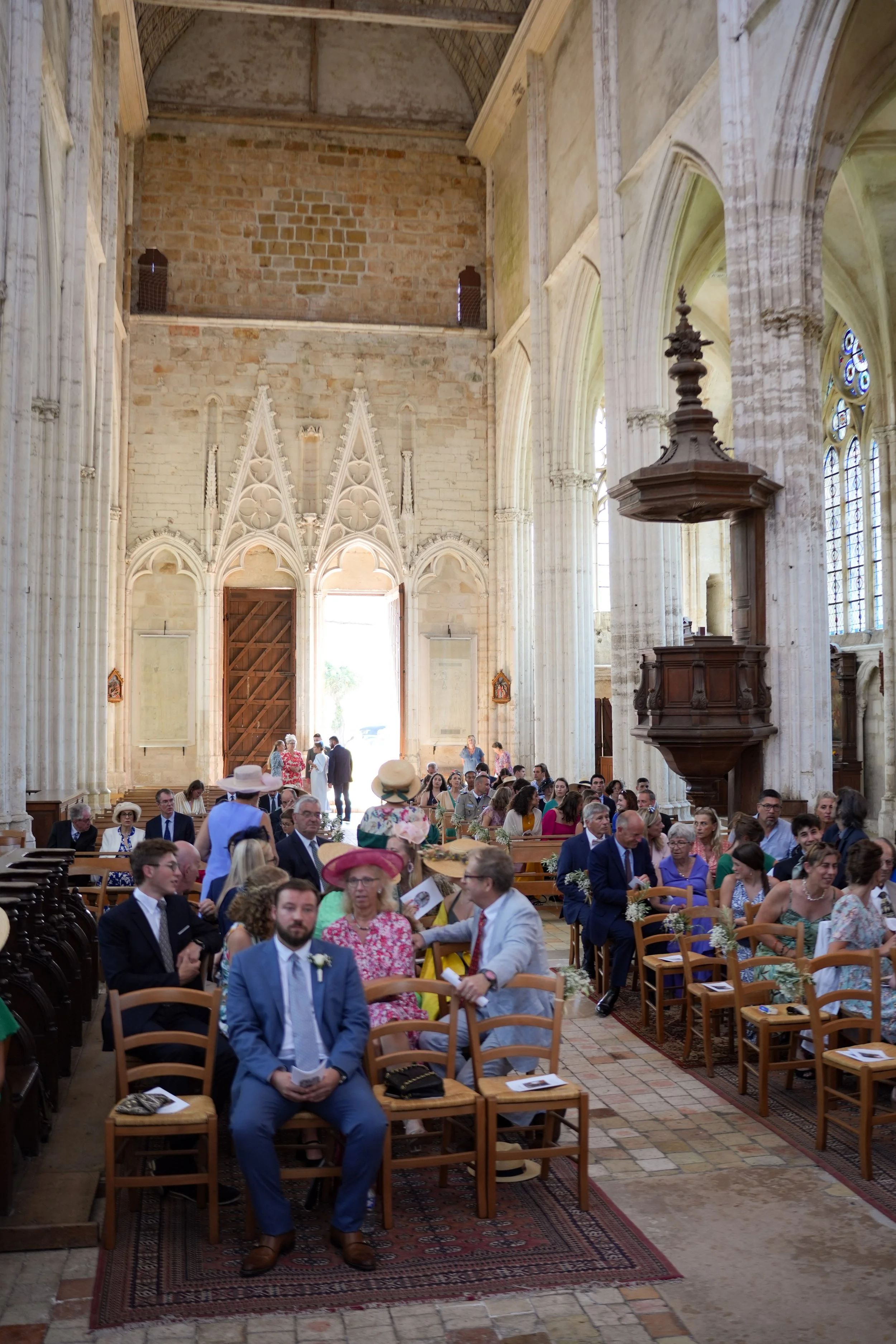 Cérémonie dans une église avec des invités assis dans des chaises en bois, portant des vêtements formels et des chapeaux, sous une architecture gothique avec de hautes fenêtres et des piliers en pierre.