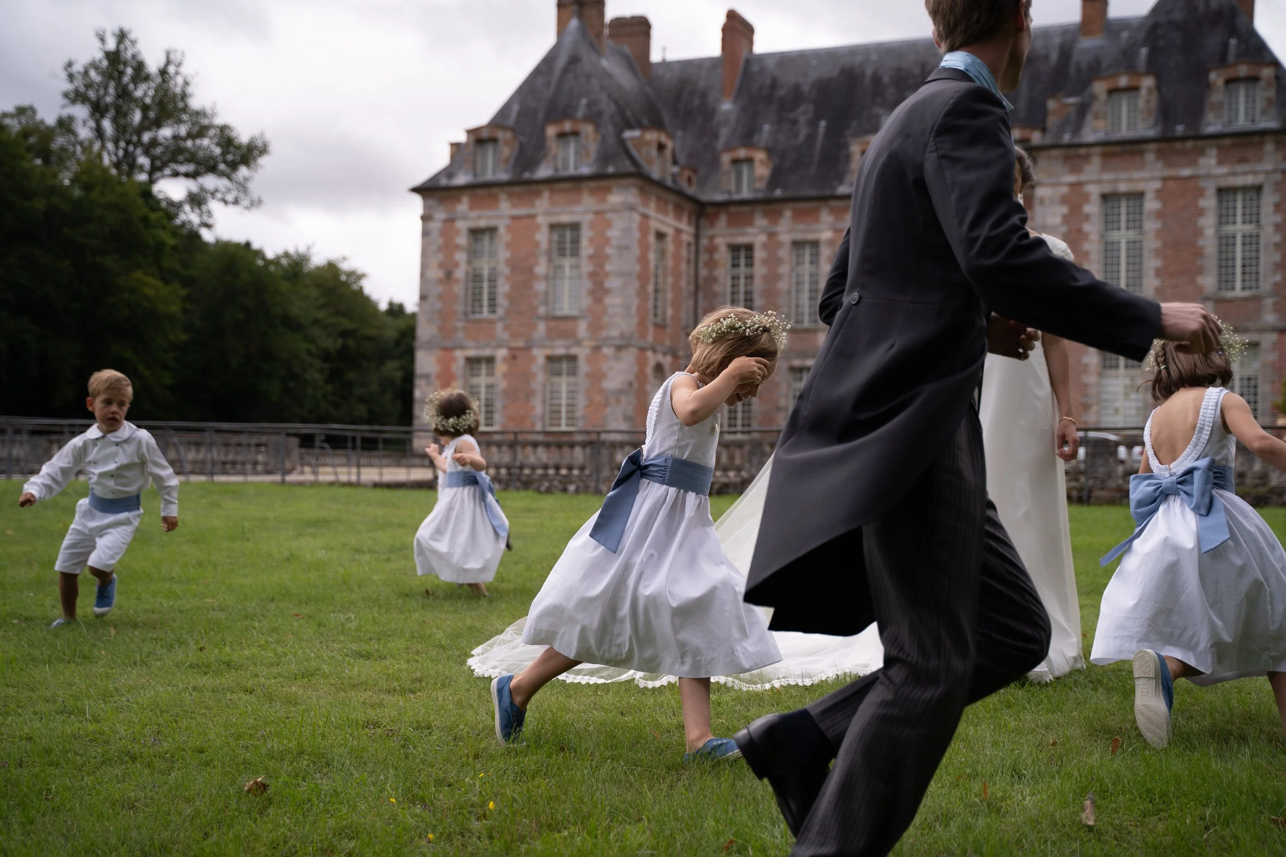 Enfant en robe blanche et couronne de fleurs courant sur la pelouse d'un mariage en extérieur, avec un château en arrière-plan.