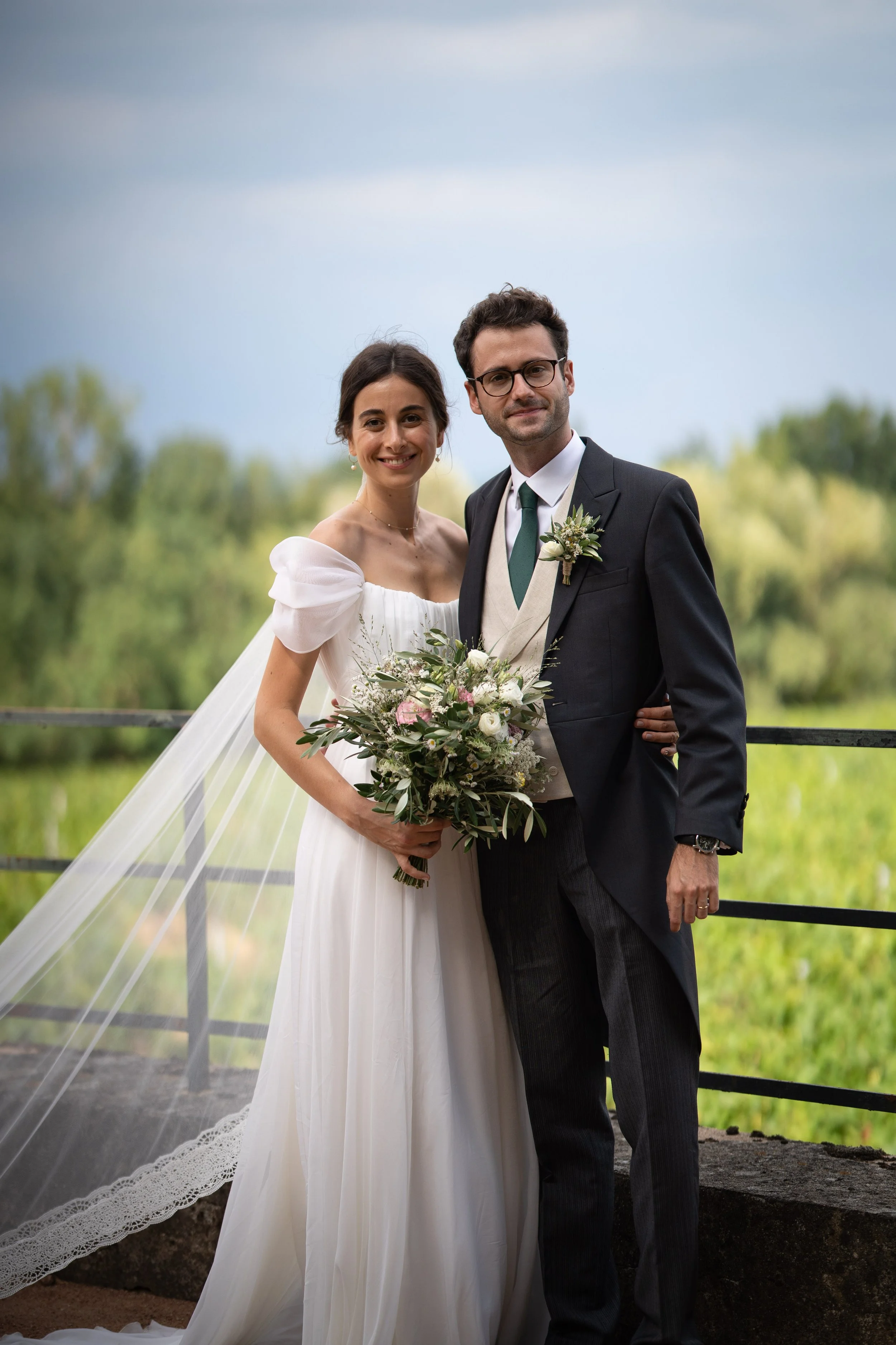 Un couple en robe de mariage et costume dans un environnement extérieur verdoyant, posant pour une photo de mariage.