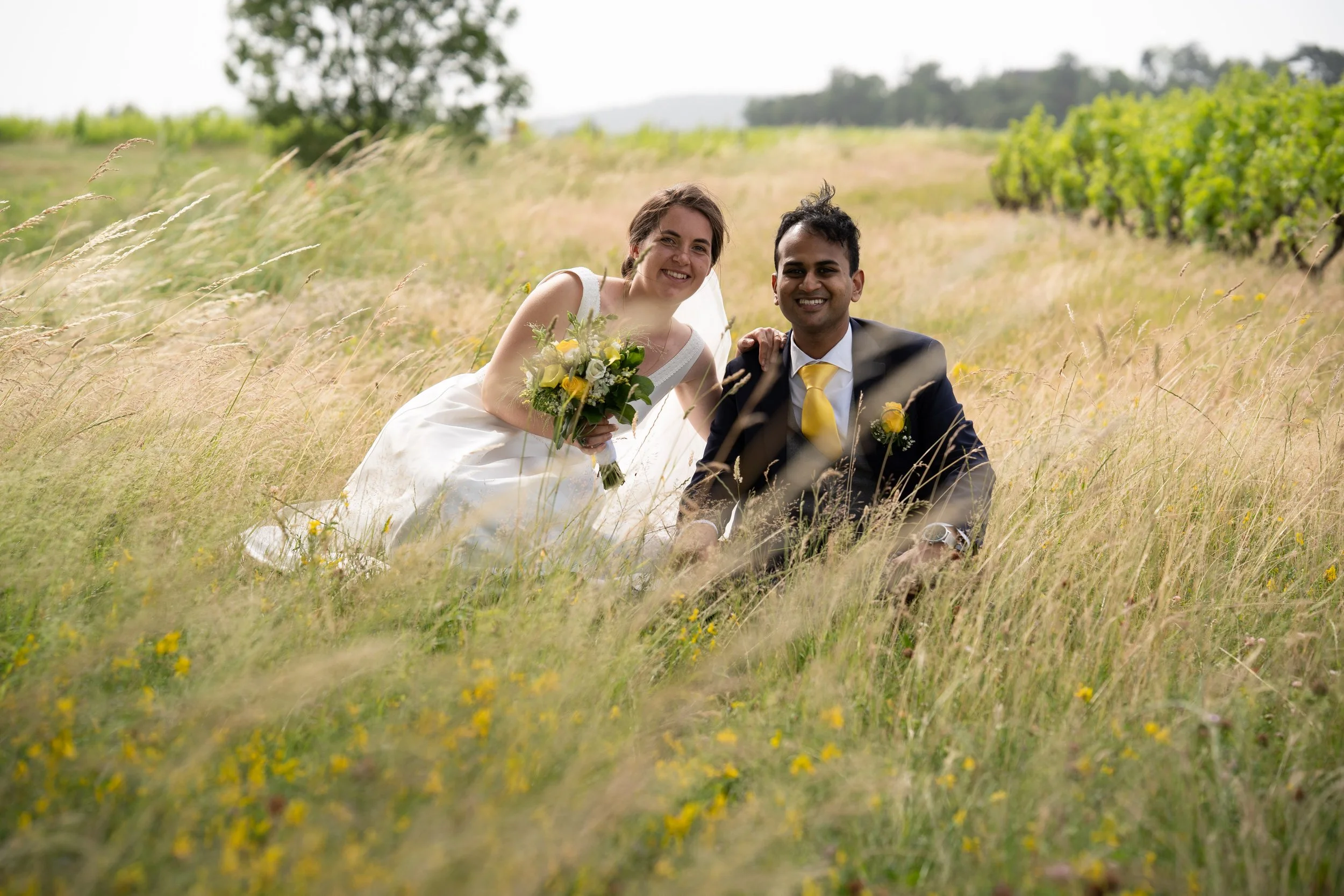 Un couple souriant en costume de mariage, assis dans un champ d'herbes hautes avec un bouquet de fleurs, dans un paysage rural avec des arbres et des collines au loin.
