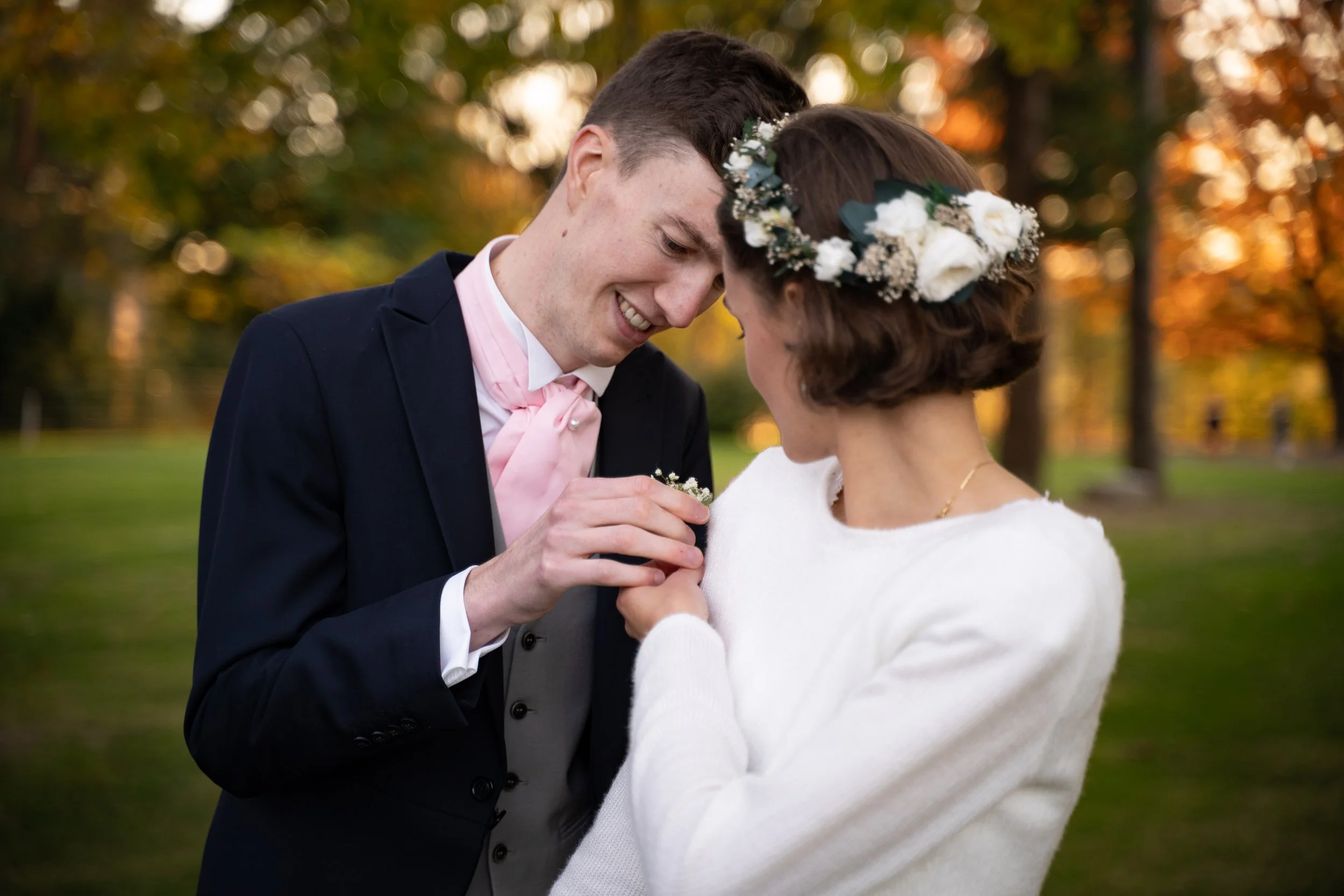 Un couple en tenue de mariage échange une bague lors d'une cérémonie en extérieur avec des arbres en arrière-plan aux couleurs d'automne.