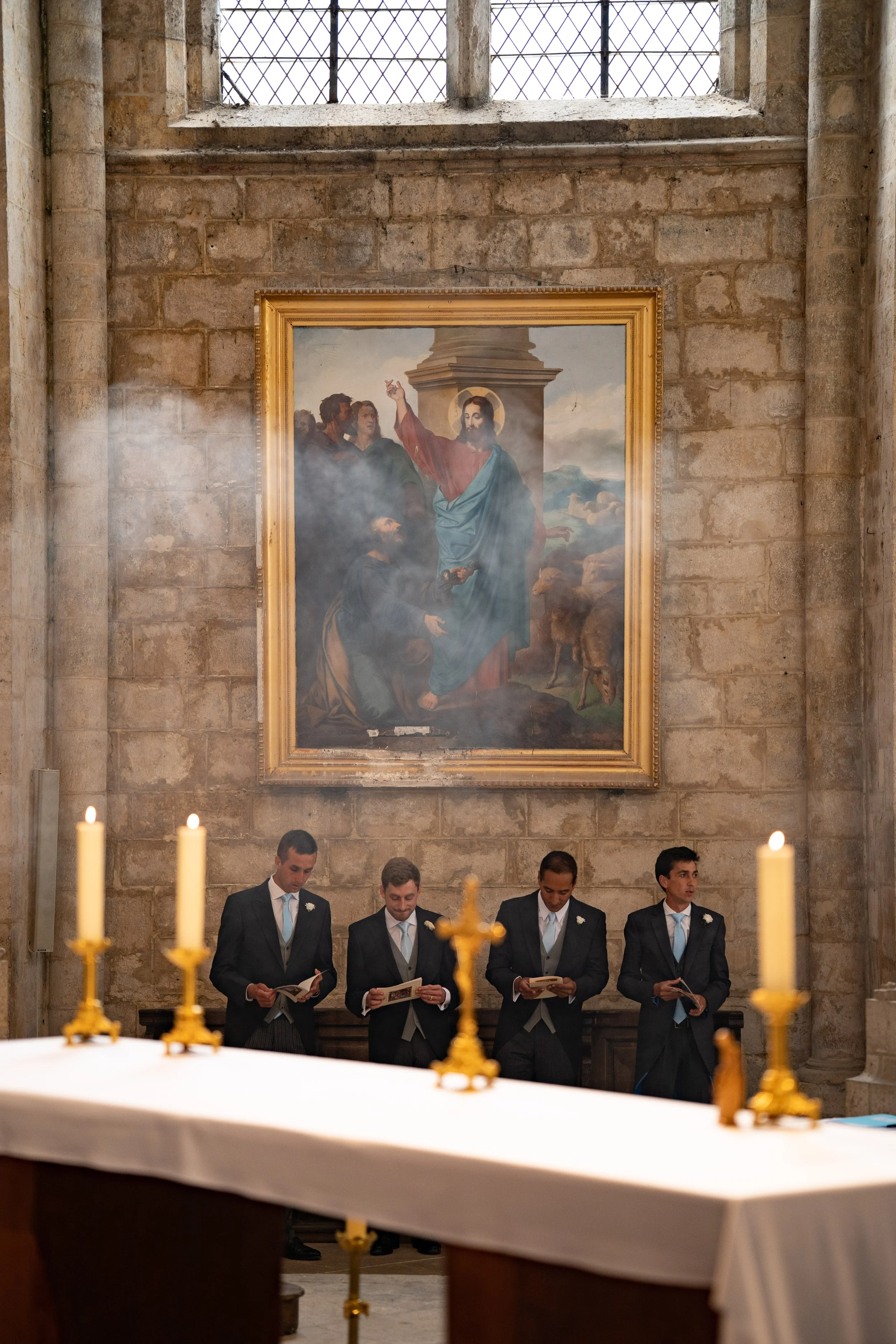 Quatre hommes en costume noir et cravate chantent ou lisent lors d'une cérémonie dans une église, sous un tableau religieux représentant Jésus avec plusieurs personnes, derrière une table avec des bougies