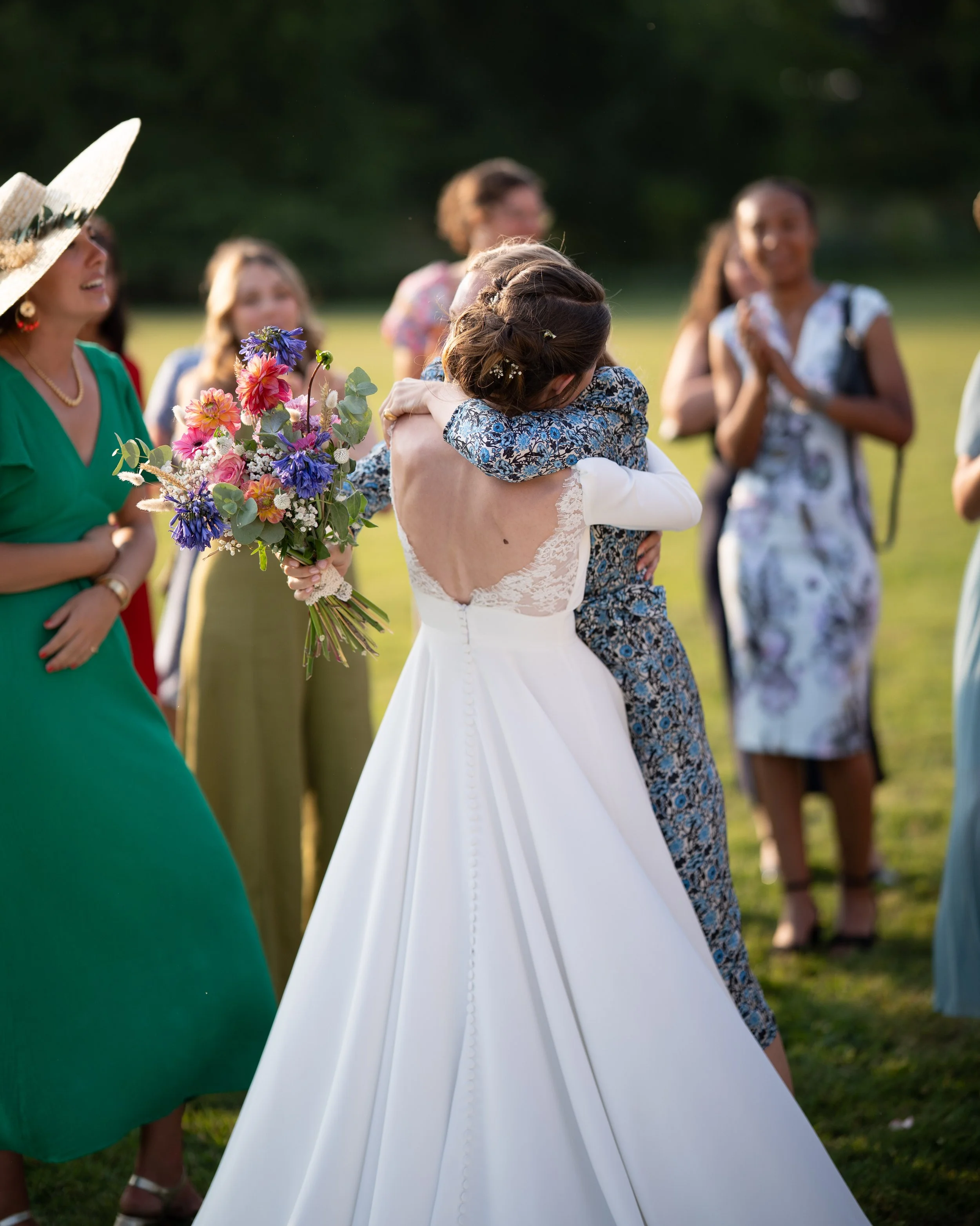 Groupe de femmes lors d'une cérémonie en plein air, avec deux femmes qui s'étreignent, une tenant un bouquet de fleurs colorées, et d'autres souriantes dans le fond.