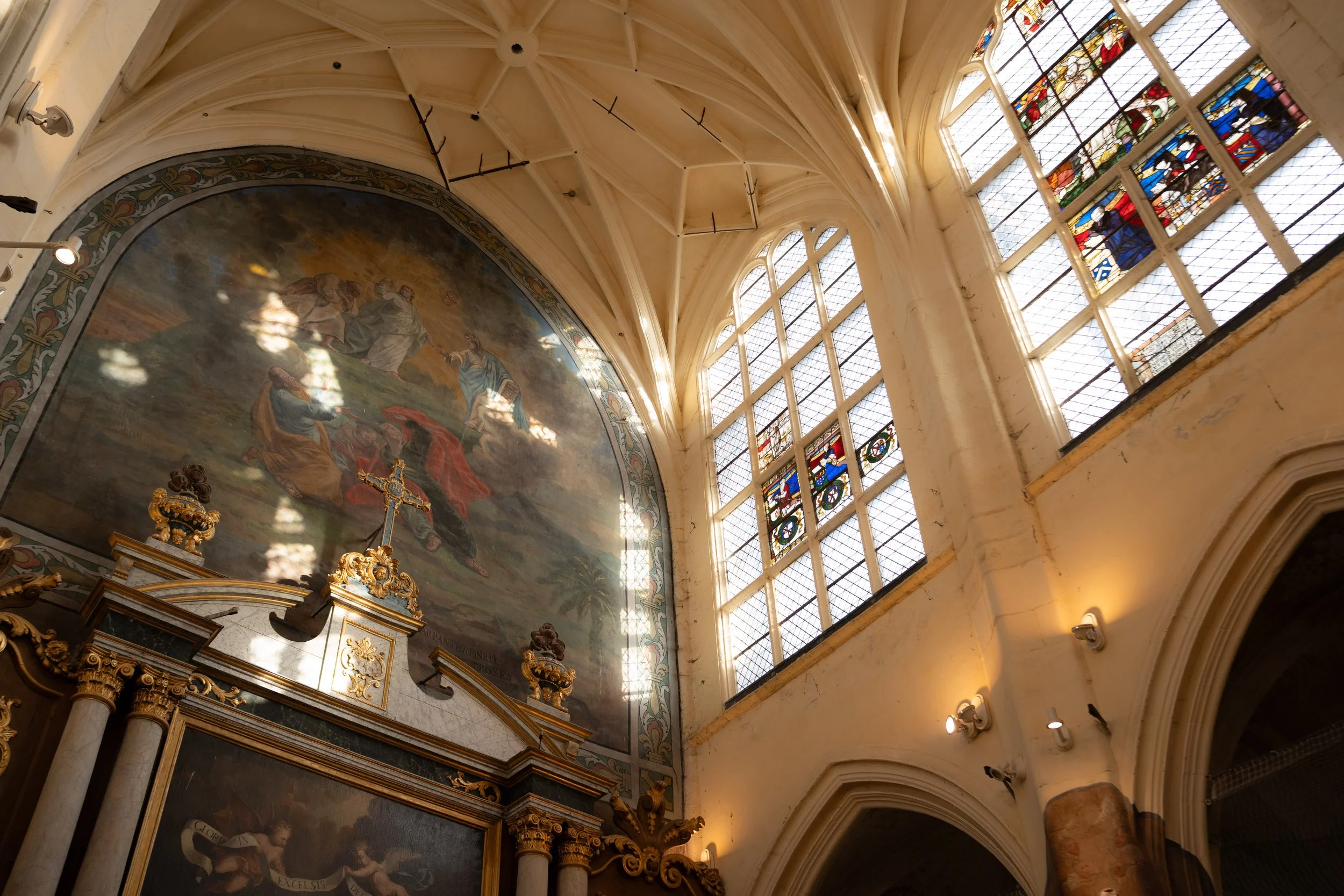 Intérieur d'une église avec des vitraux colorés et un plafond en voûte, mettant en valeur un grand tableau religieux et des colonnes en marbre avec des détails dorés.