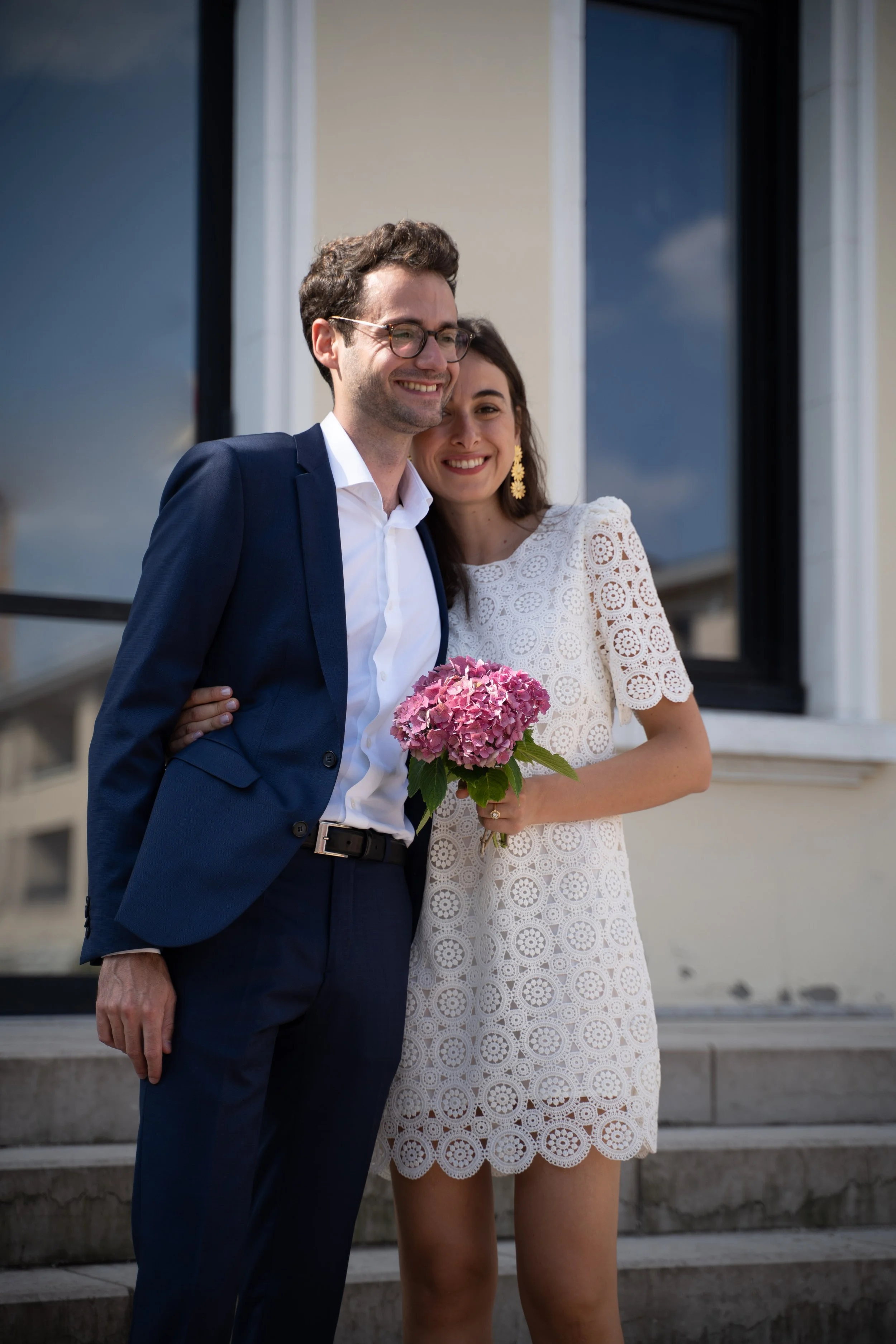 Un couple souriant se tenant par la taille devant un bâtiment, la femme tenant un bouquet de hortensias roses. La femme porte une robe blanche avec des motifs circulaires, et des boucles d'oreilles dorées. L'homme porte un costume bleu foncé avec une