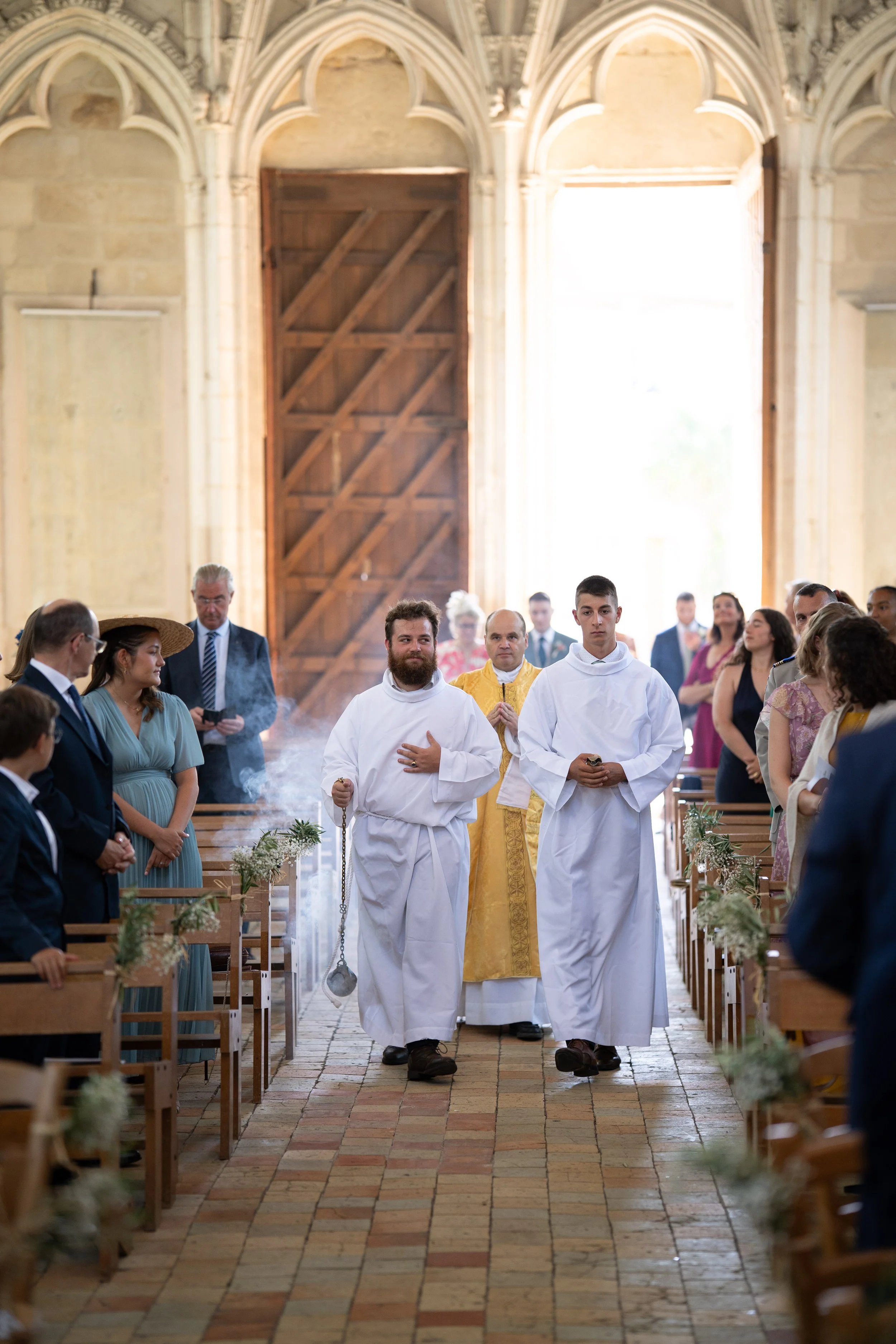 Une cérémonie religieuse dans une église avec des personnes assises et deux jeunes prêtres marchant dans l'allée centrale.