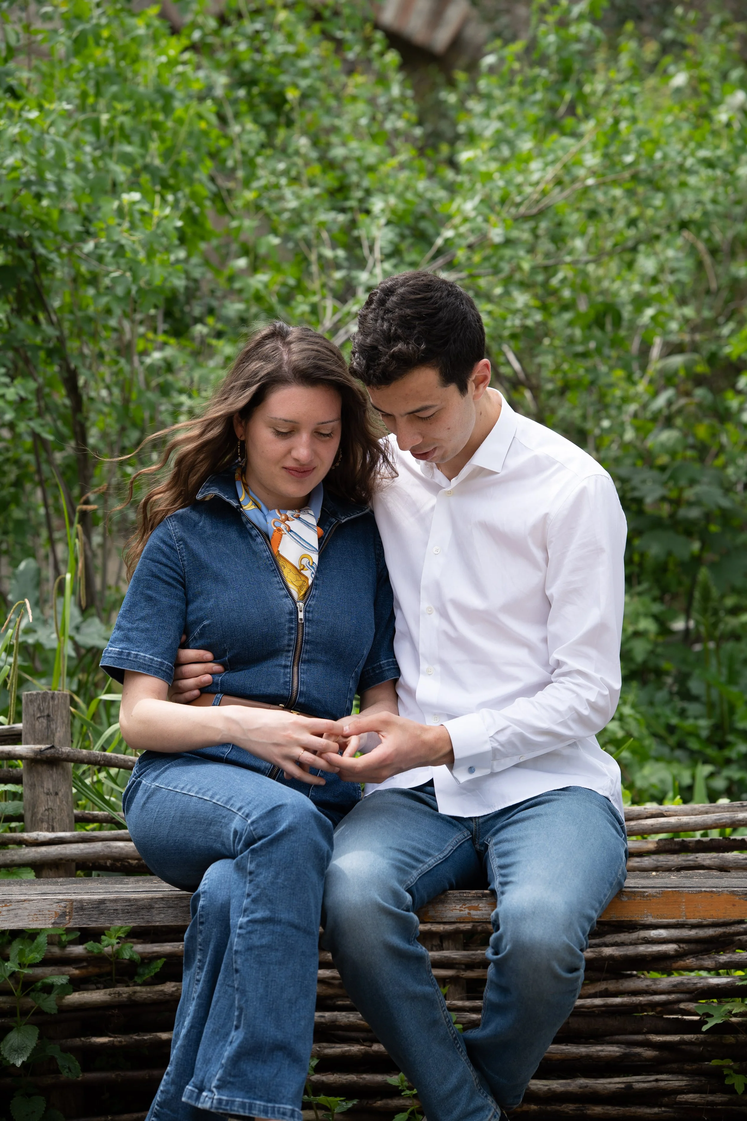 Un couple assis sur un banc en bois dans un jardin, regardant un téléphone portable ensemble, entouré de verdure.