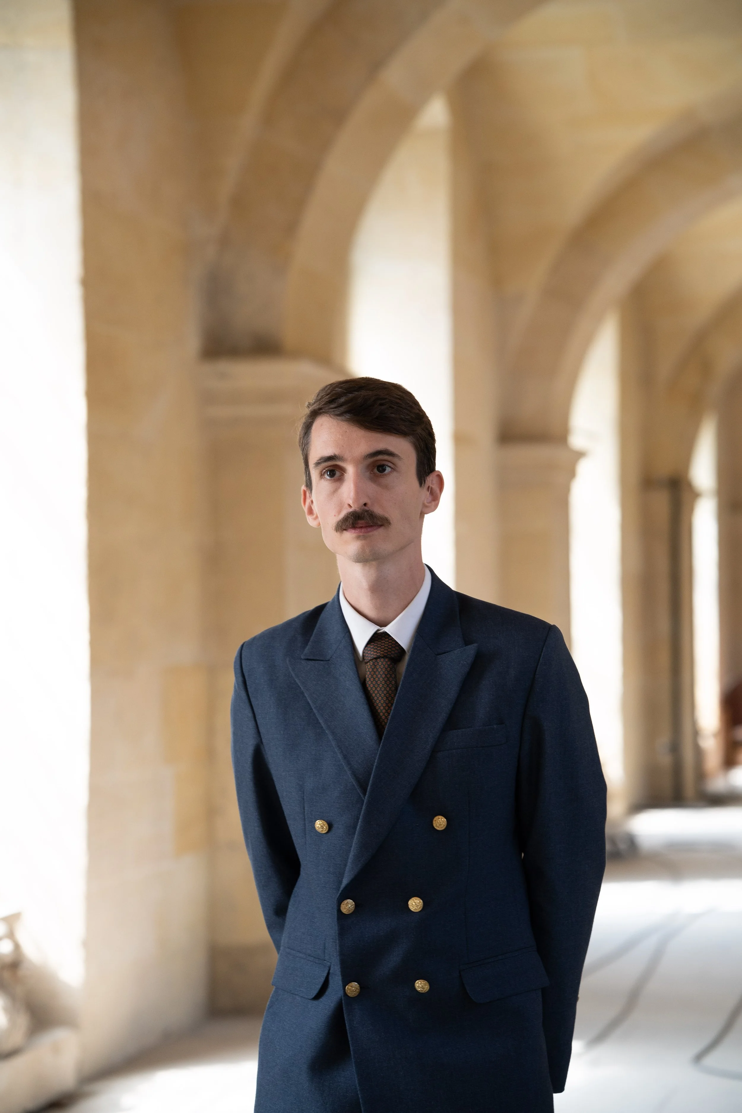 Un homme en uniforme bleu marine avec des boutons dorés, portant une cravate, debout dans un couloir avec des arches en pierre beige.