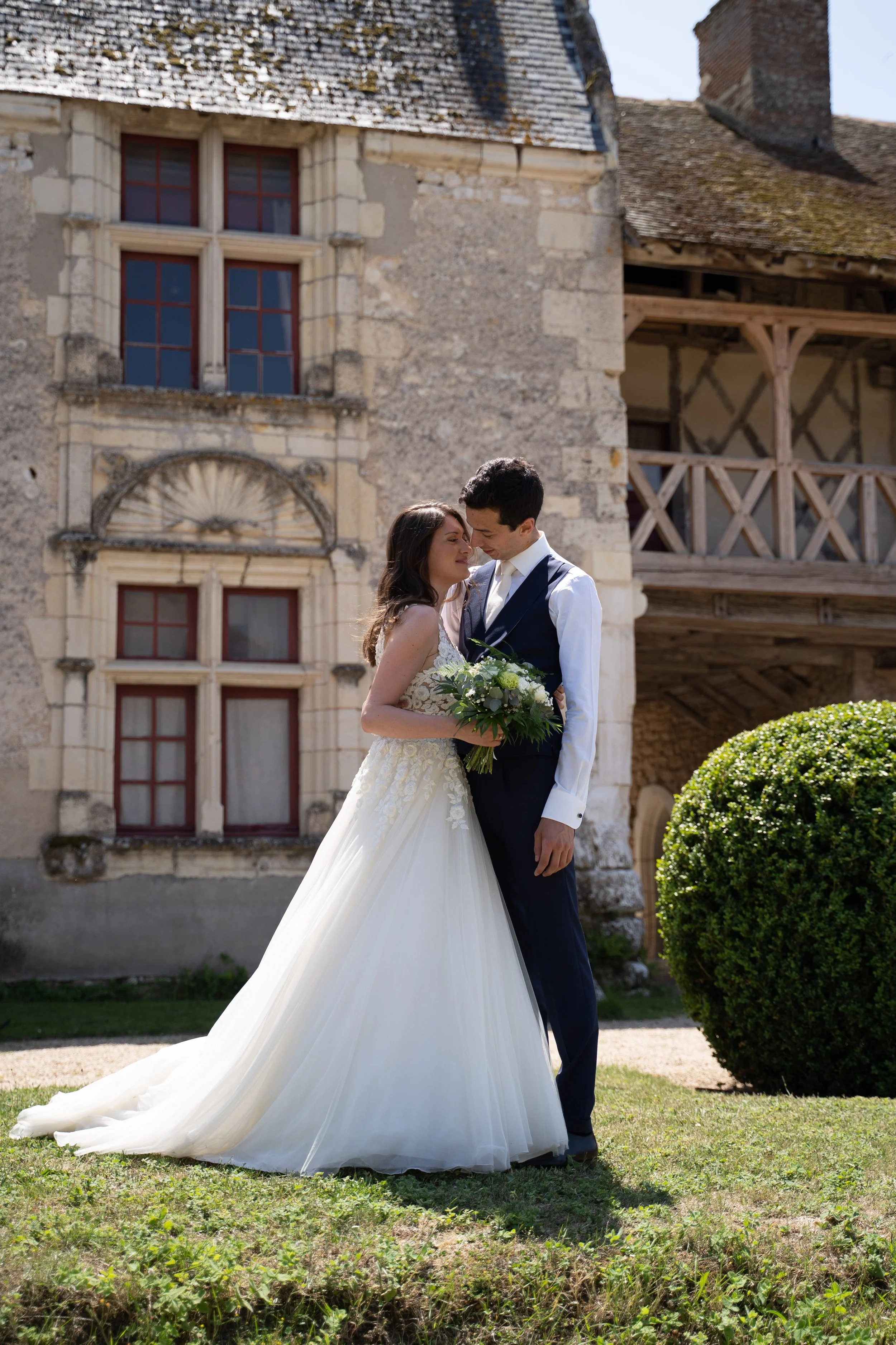 Un couple marié, la femme en robe de mariée blanche longue et le homme en costume sombre, se tiennent près d'une vieille bâtisse en pierre avec fenêtres à volets rouges et balcon en bois, lors d'une photographie de mariage en extérieur.