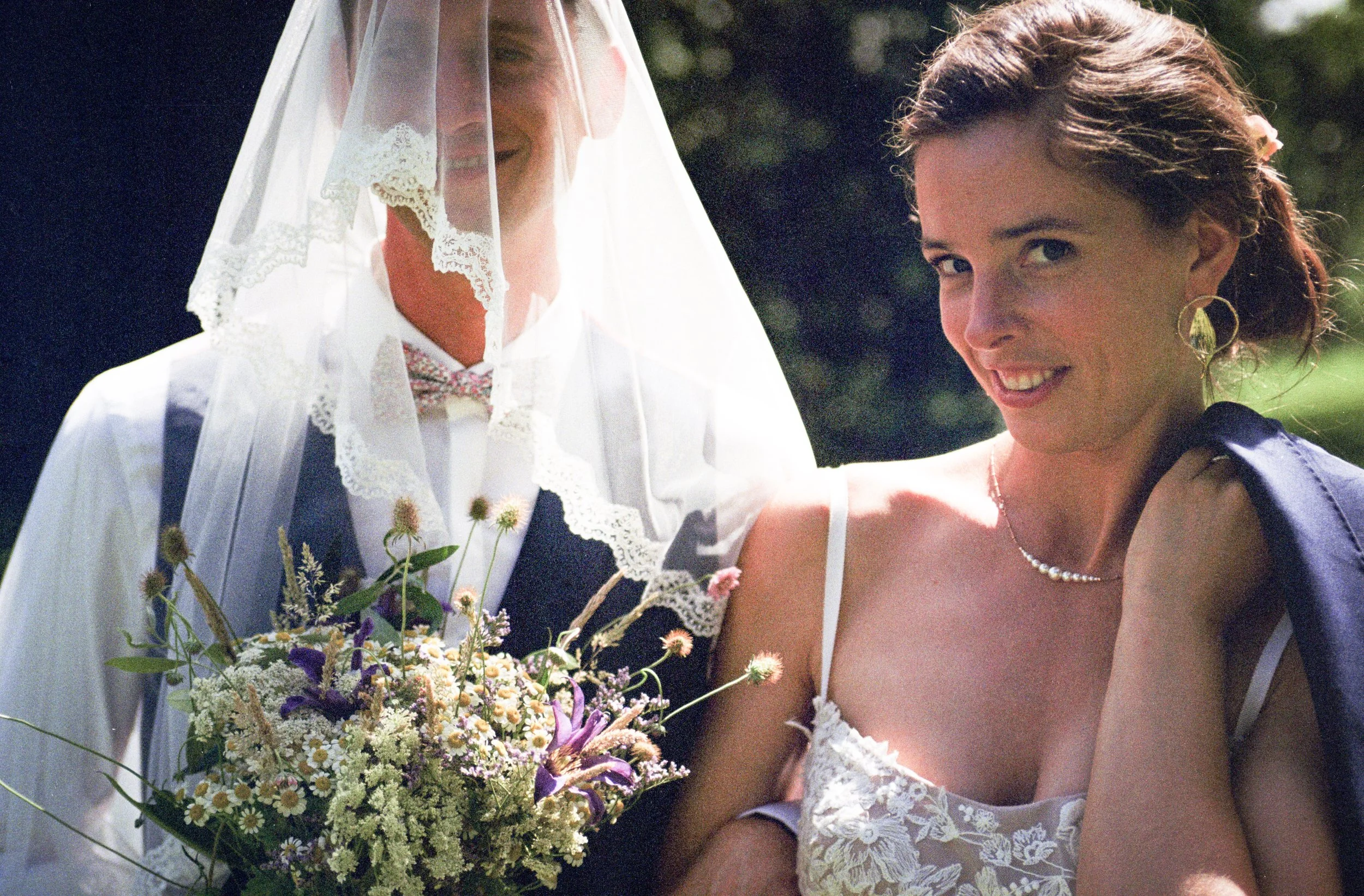 Une femme en robe de mariée sourit à côté d'une autre femme en robe de soirée qui lui offre un bouquet de fleurs lors d'un mariage en extérieur.
