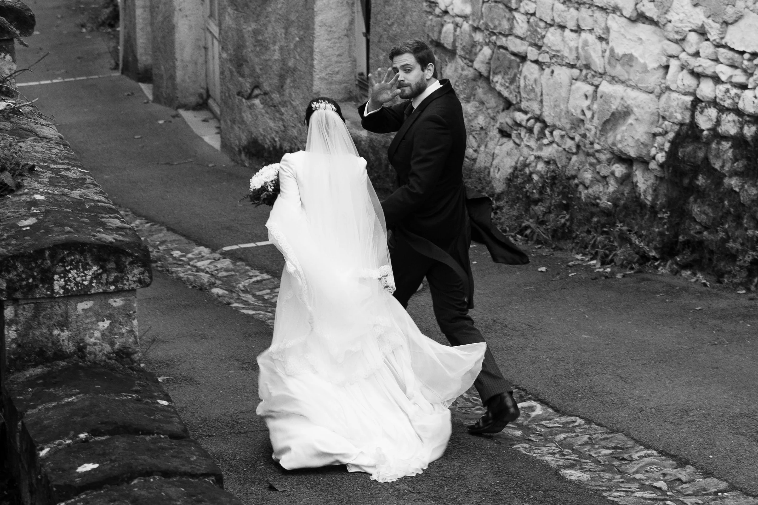 Un homme en costume noir, probablement un marié, danse avec une femme en robe de mariée dans une ruelle pavée en pierre, entourés de murs en pierre. La scène est en noir et blanc.