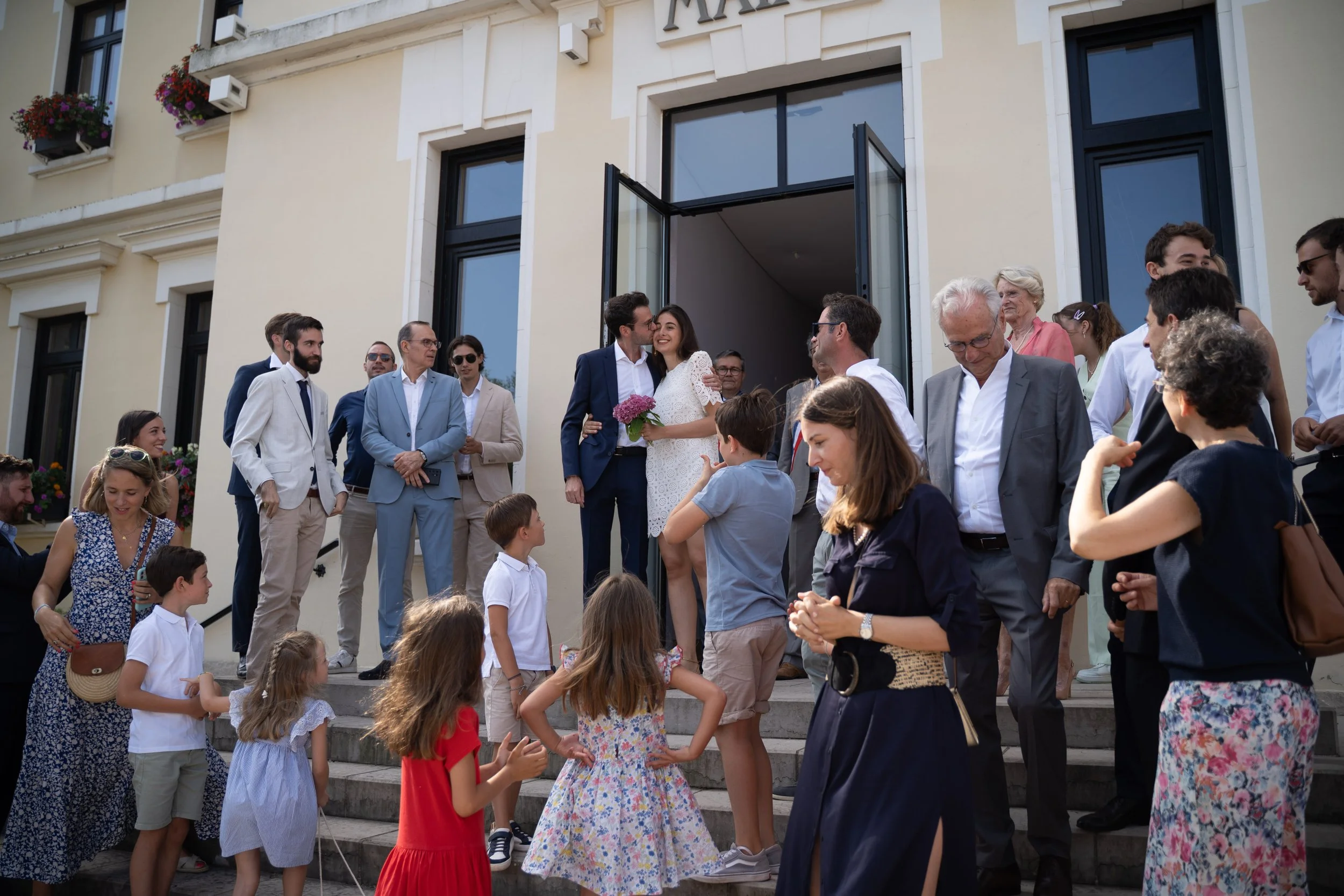Groupe de personnes, adultes et enfants, réunis devant un bâtiment pour une célébration ou un rassemblement social, avec des personnes qui discutent, sourient et se saluent.