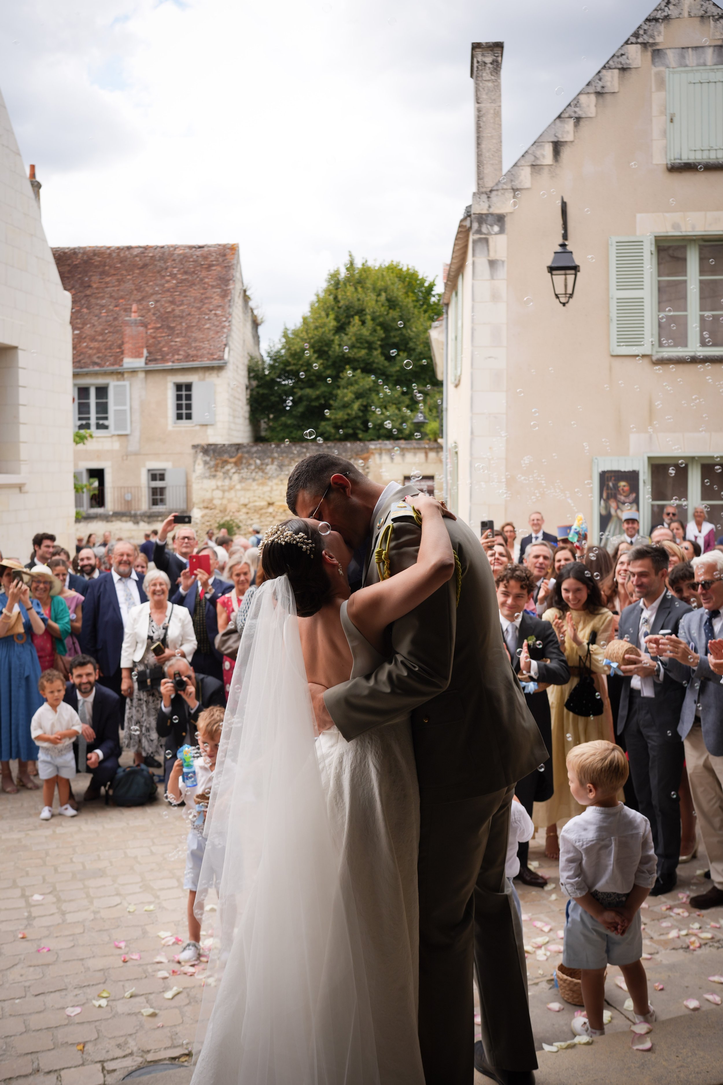 Un couple de mariés s'embrassent lors de leur mariage dans une cour médiévale, entourés d'invités, avec des maisons anciennes en arrière-plan.