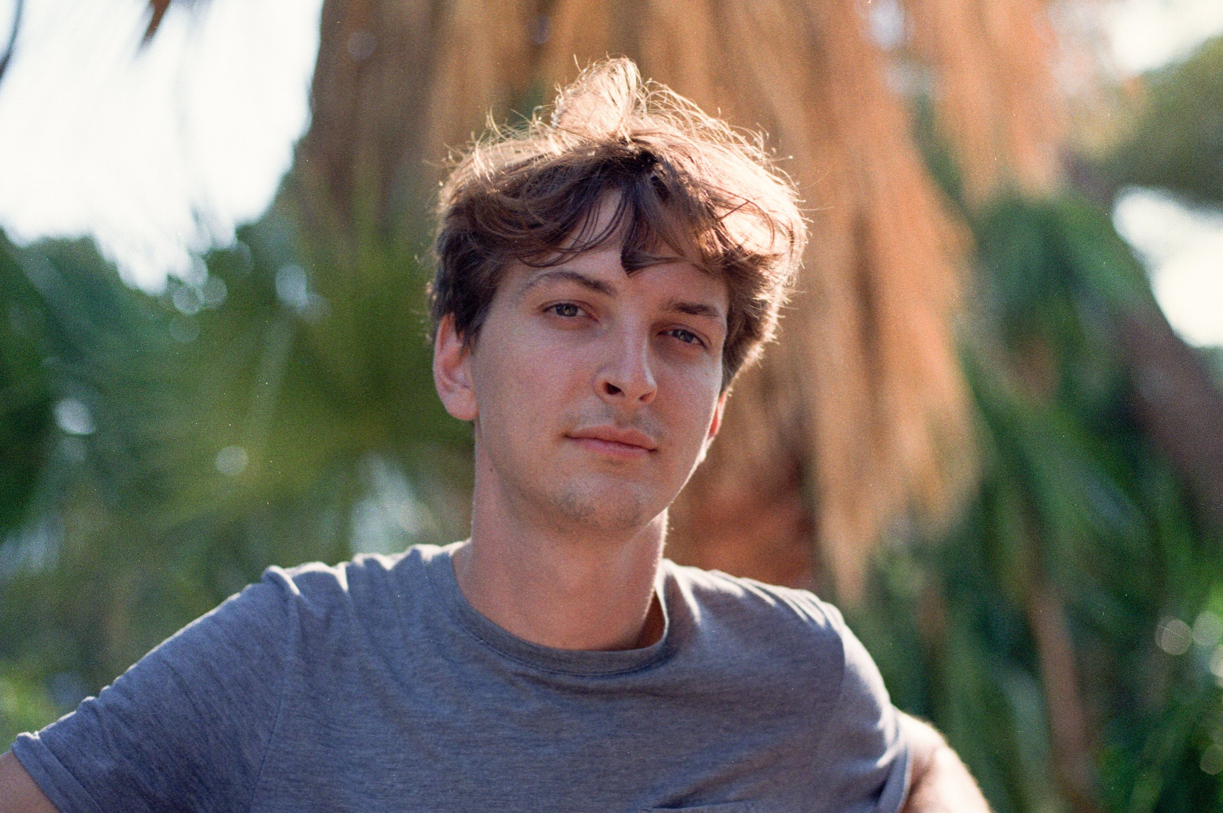 Jeune homme avec des cheveux bouclés et des yeux bleus, portant un t-shirt gris, en extérieur avec un arbre en arrière-plan.