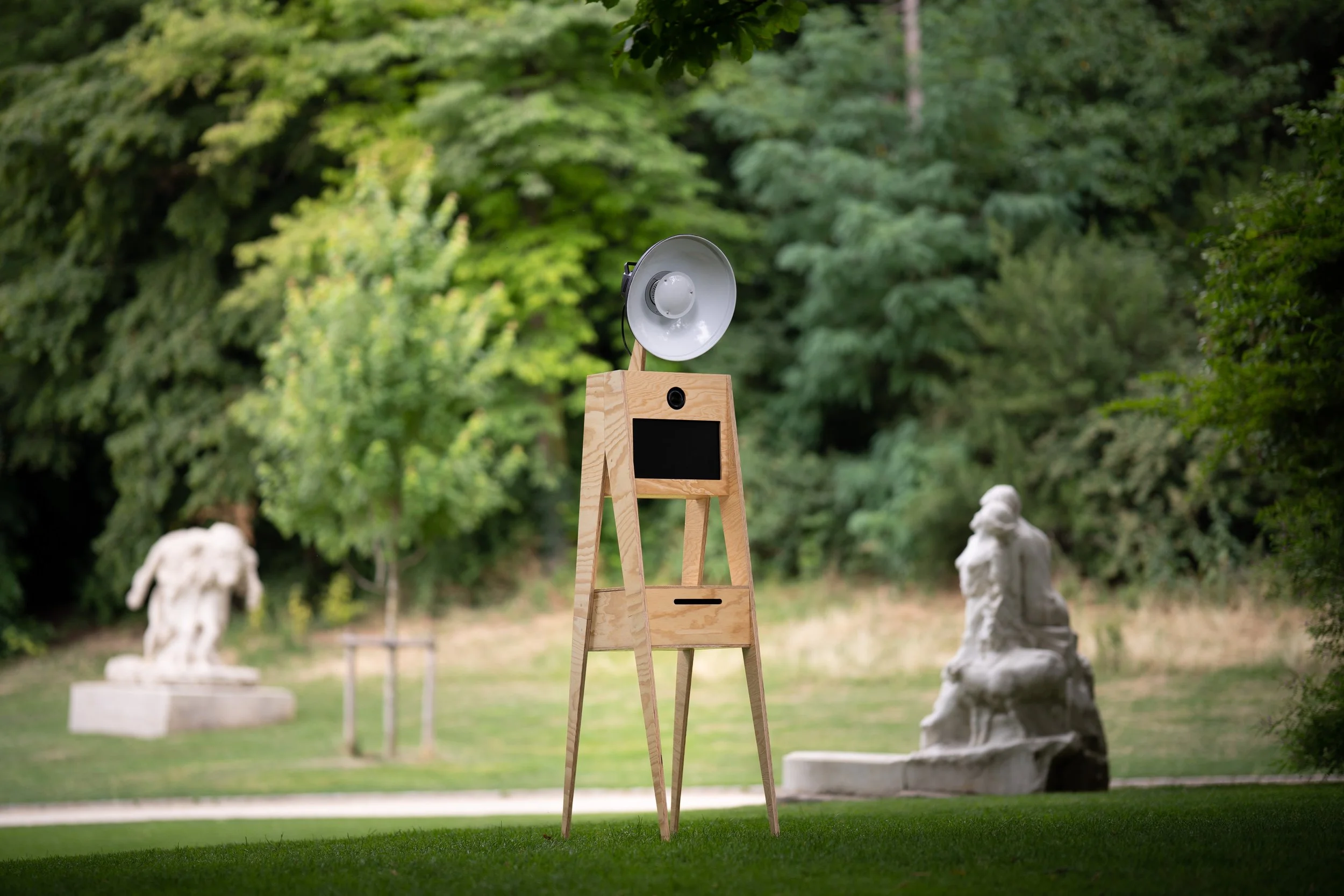 Une sculpture de style classique sur une base en pierre dans un jardin, avec un arbre feuillu en arrière-plan.