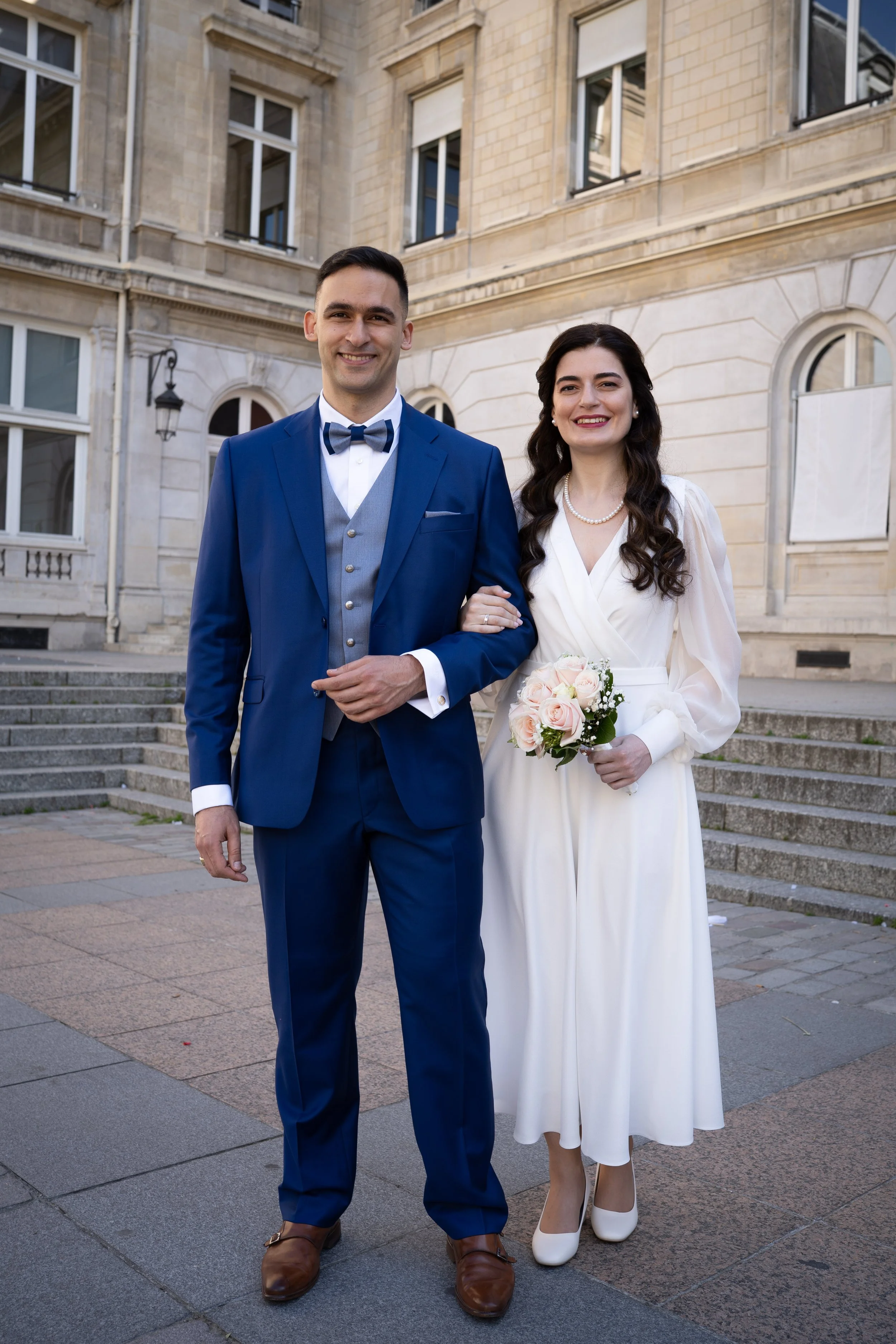Un couple en costume de mariage posant devant un bâtiment en pierre. L'homme porte un costume bleu avec une cravate à motif, et la femme porte une robe blanche avec un bouquet de roses pâles.