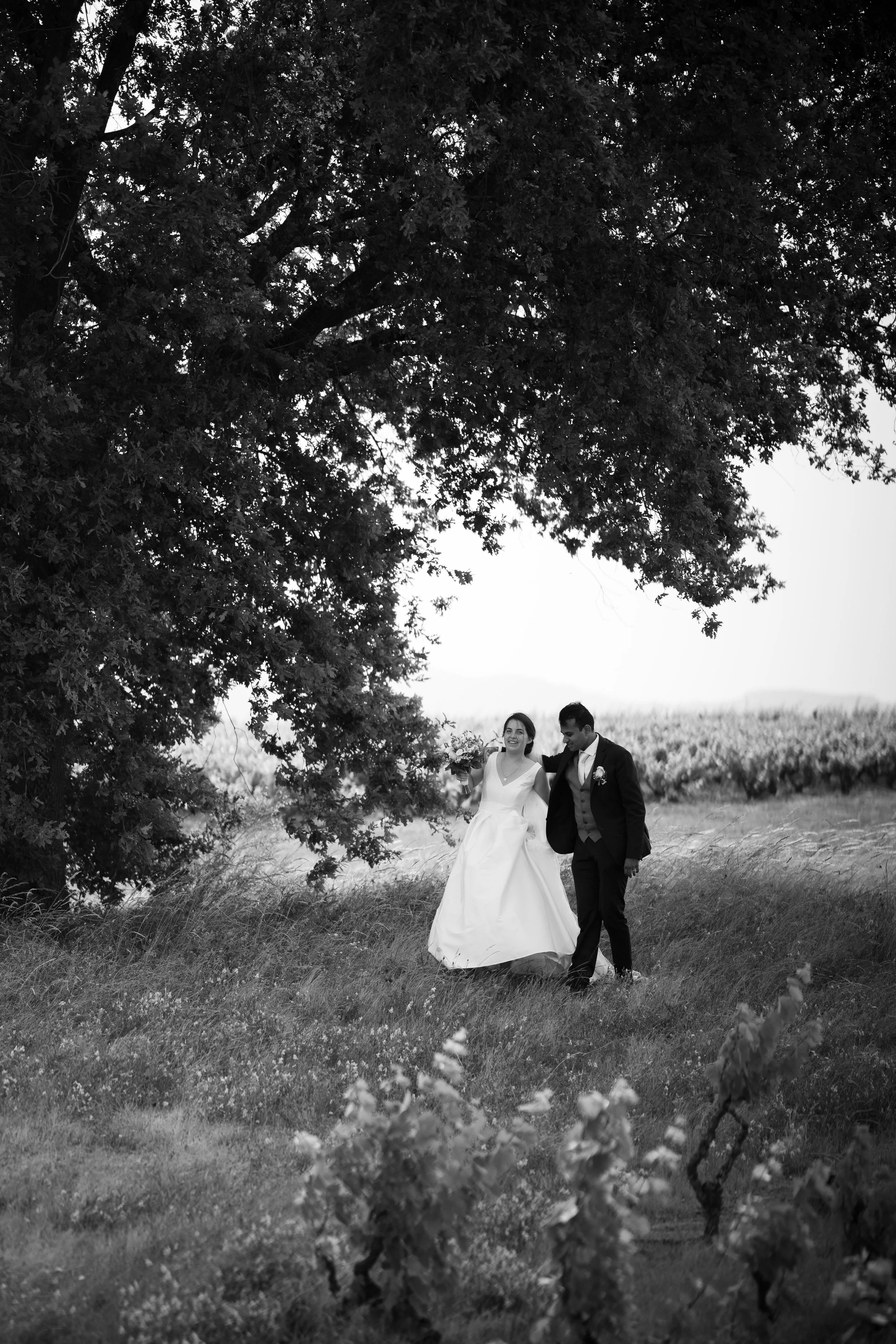 Un couple de mariés marchant dans un champ avec beaucoup d'arbres, en noir et blanc.