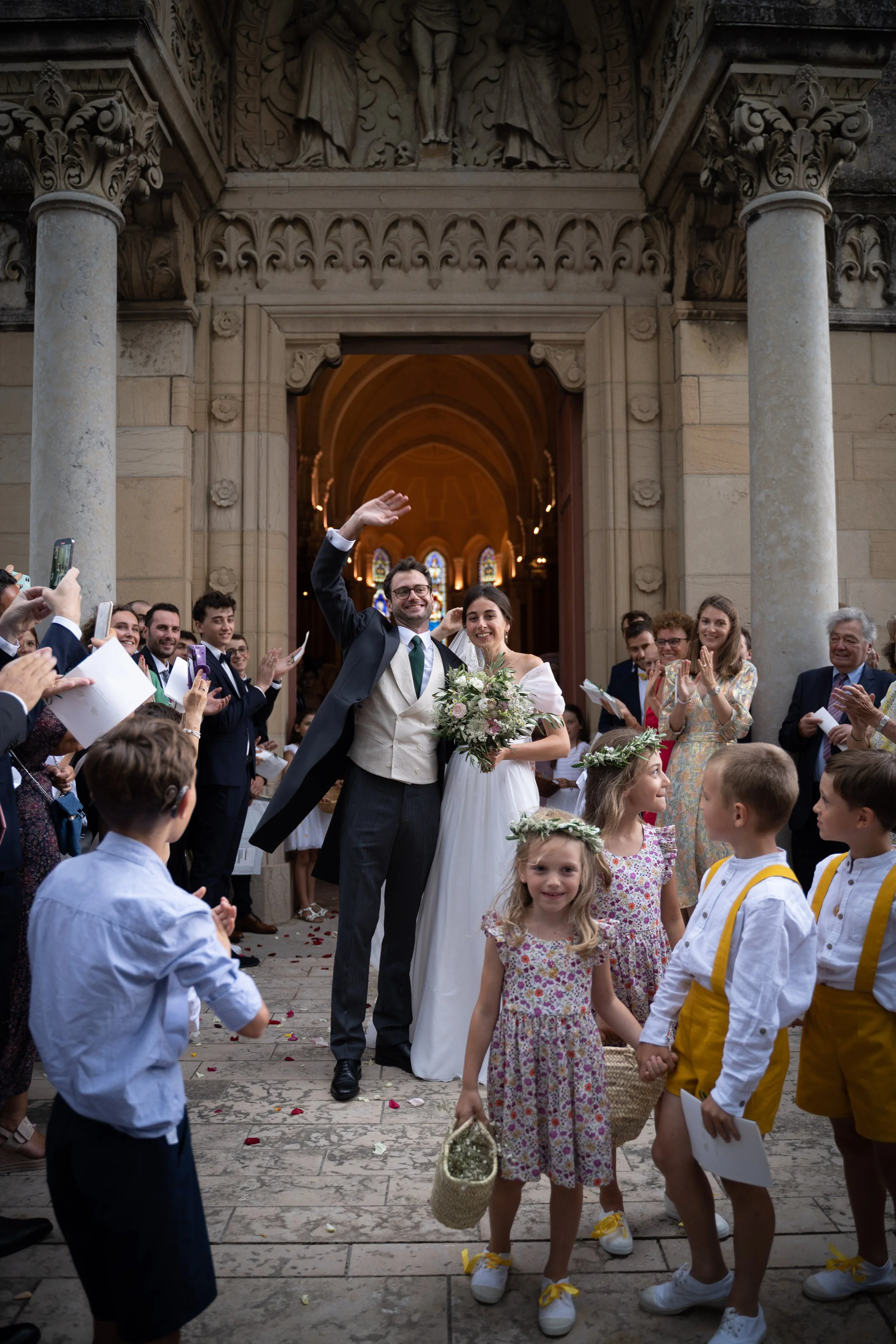 Un couple de mariés souriants sortant d'une église après leur mariage, entourés de familles et amis. La scène est joyeuse avec des personnes qui prennent des photos et lancent des pétales.