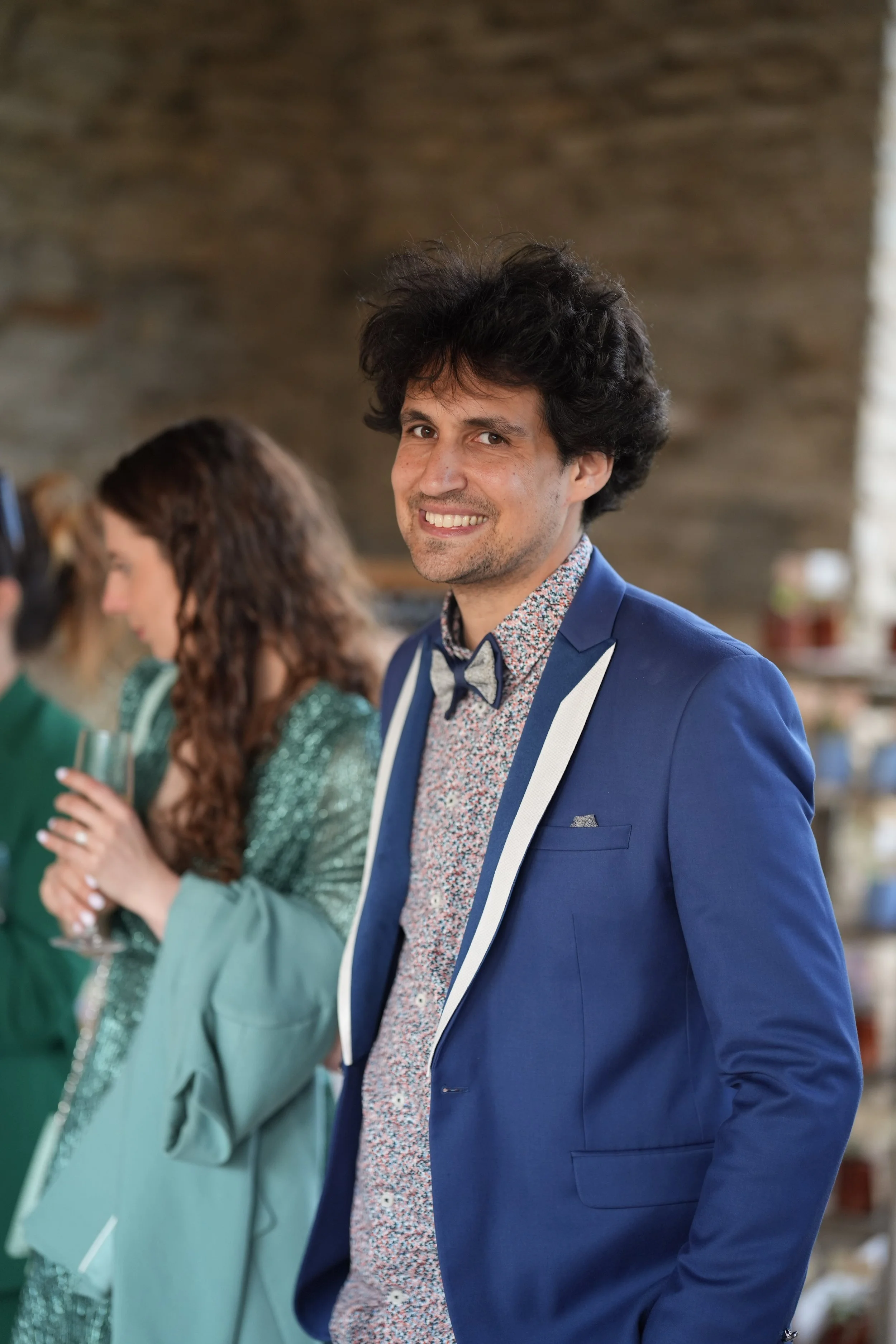 Un homme en costume bleu avec une chemise à motifs colorés sourit à la caméra, en arrière-plan, une femme avec des cheveux bouclés et une robe verte se tient avec un verre à la main, dans une ambiance chaleureuse.