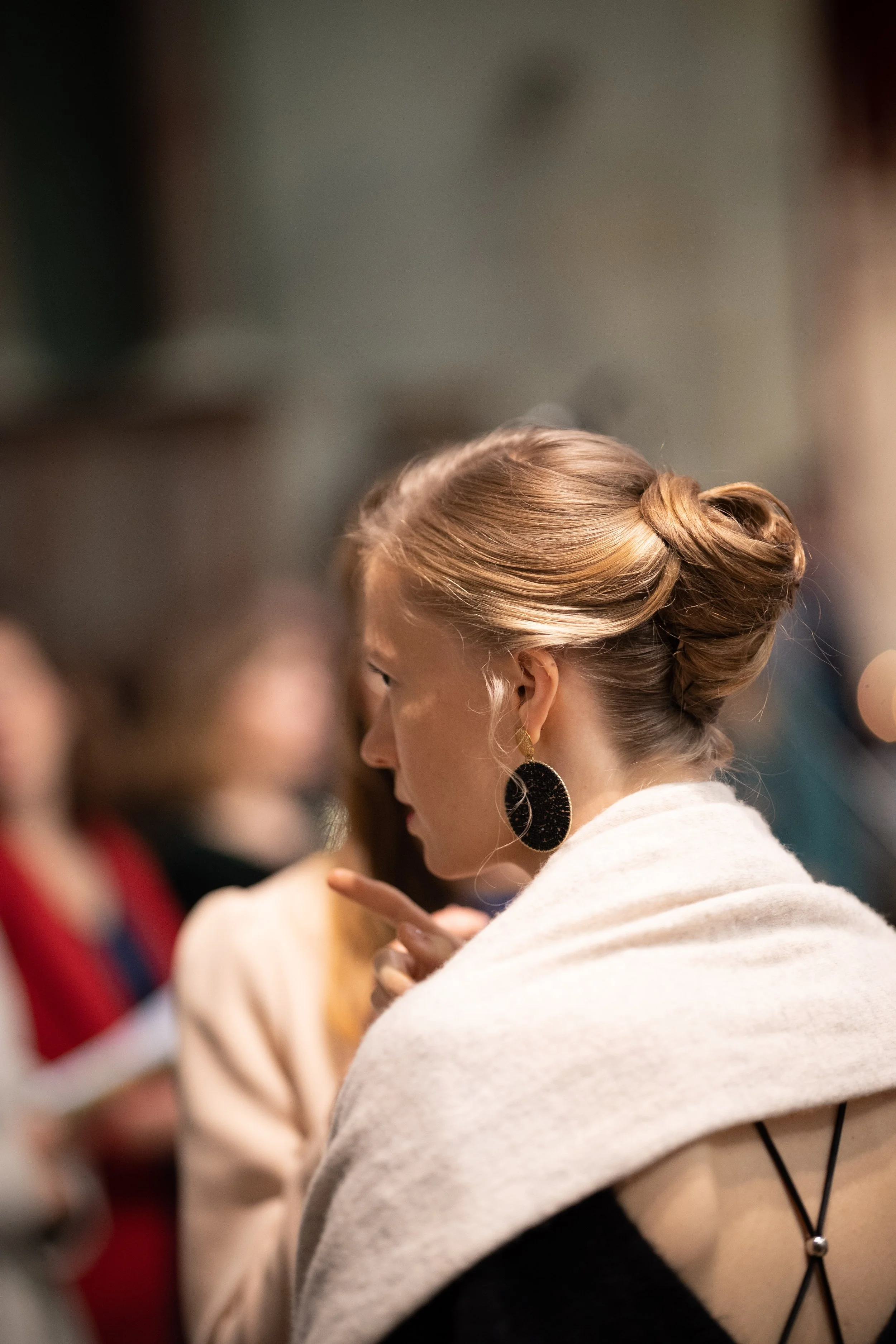 Femme aux cheveux roux coiffés en chignon, portant de grandes boucles d'oreilles noires, vêtue d'une robe beige à manches longues, assise lors d'un événement social