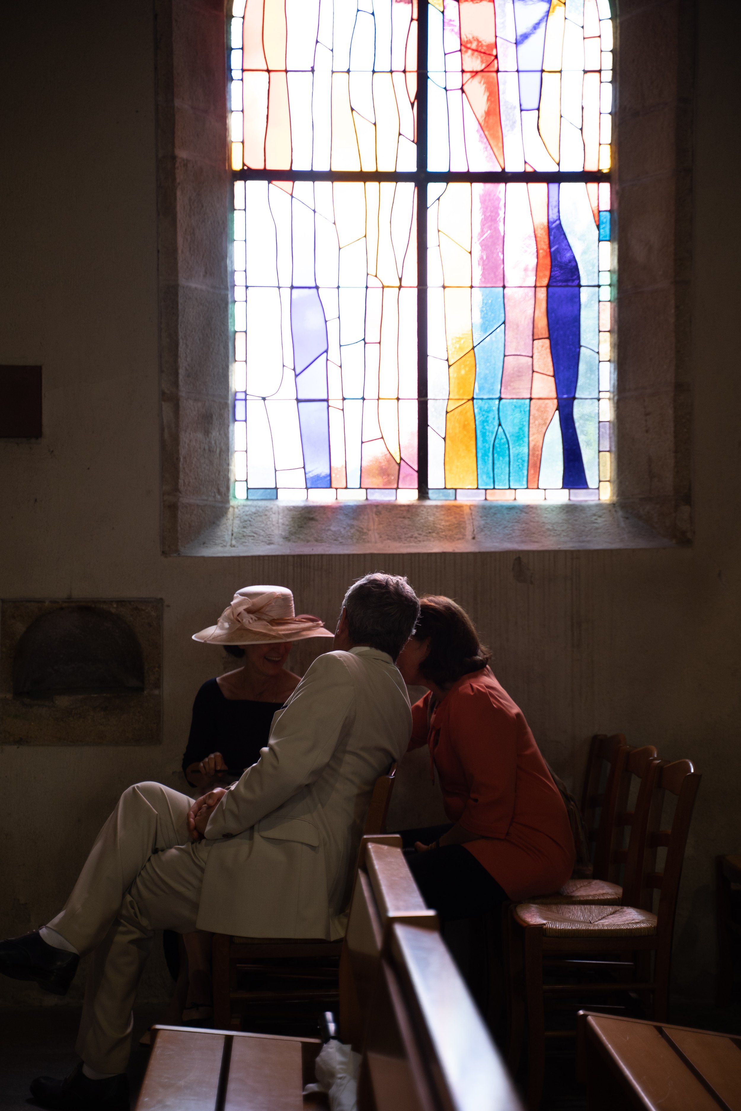 Une fenêtre en vitrail coloré lumière, avec trois personnes assises en train de discuter dans une pièce sombre. L'atmosphère est calme et intime.