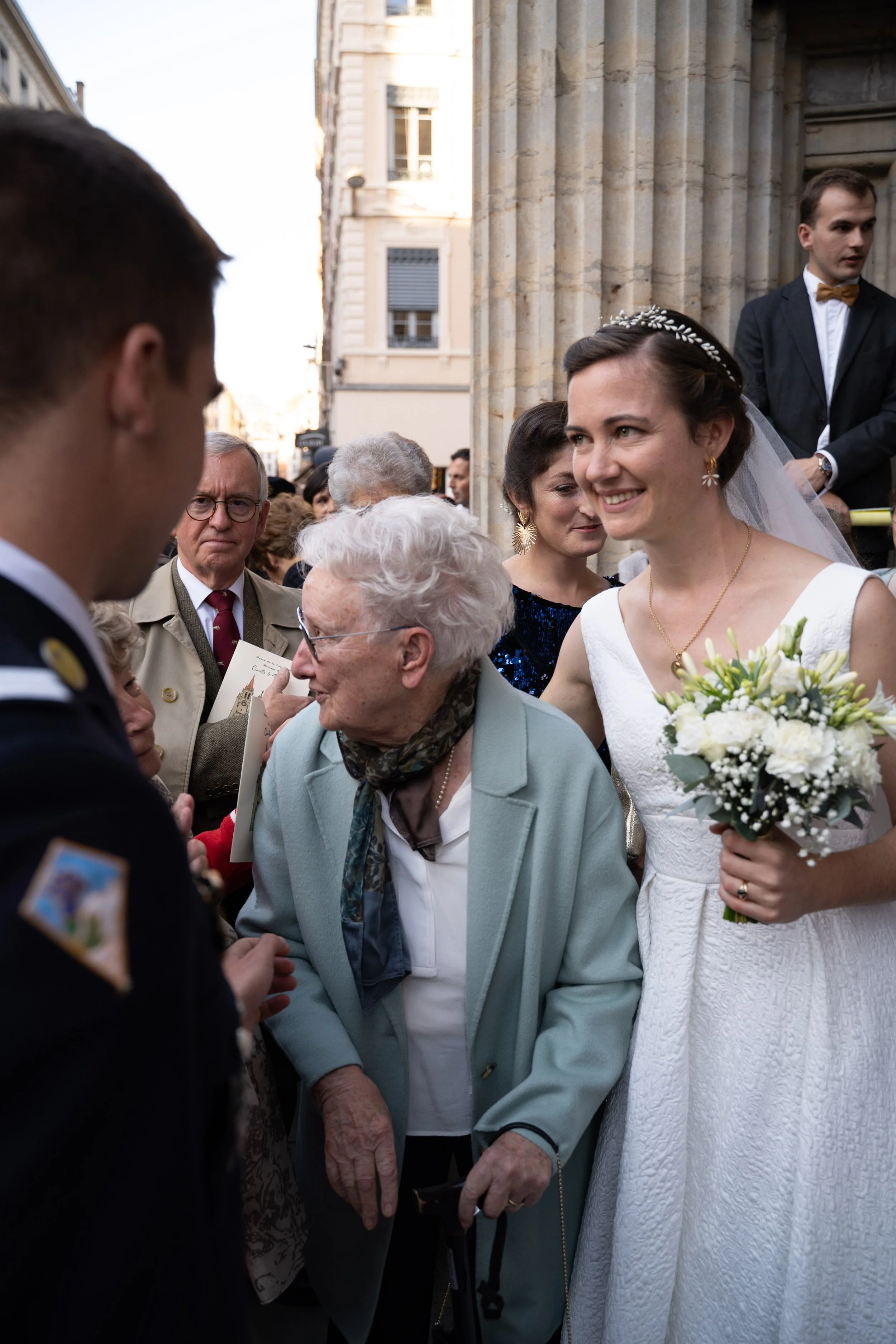 Cérémonie de mariage avec la mariée souriante, tenant un bouquet de fleurs blanches, entourée de plusieurs personnes devant une église en arrière-plan.