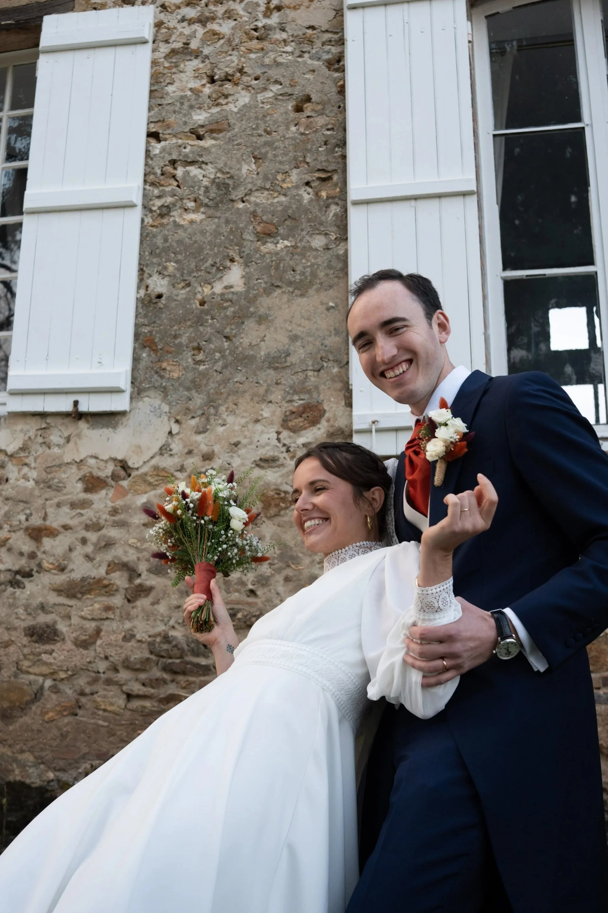 Un couple de mariés souriant, la femme portant une robe blanche et tenant un bouquet de fleurs, l'homme en costume sombre avec une boutonnière, en célébration près d'une vieille bâtisse en pierre avec volets blancs.