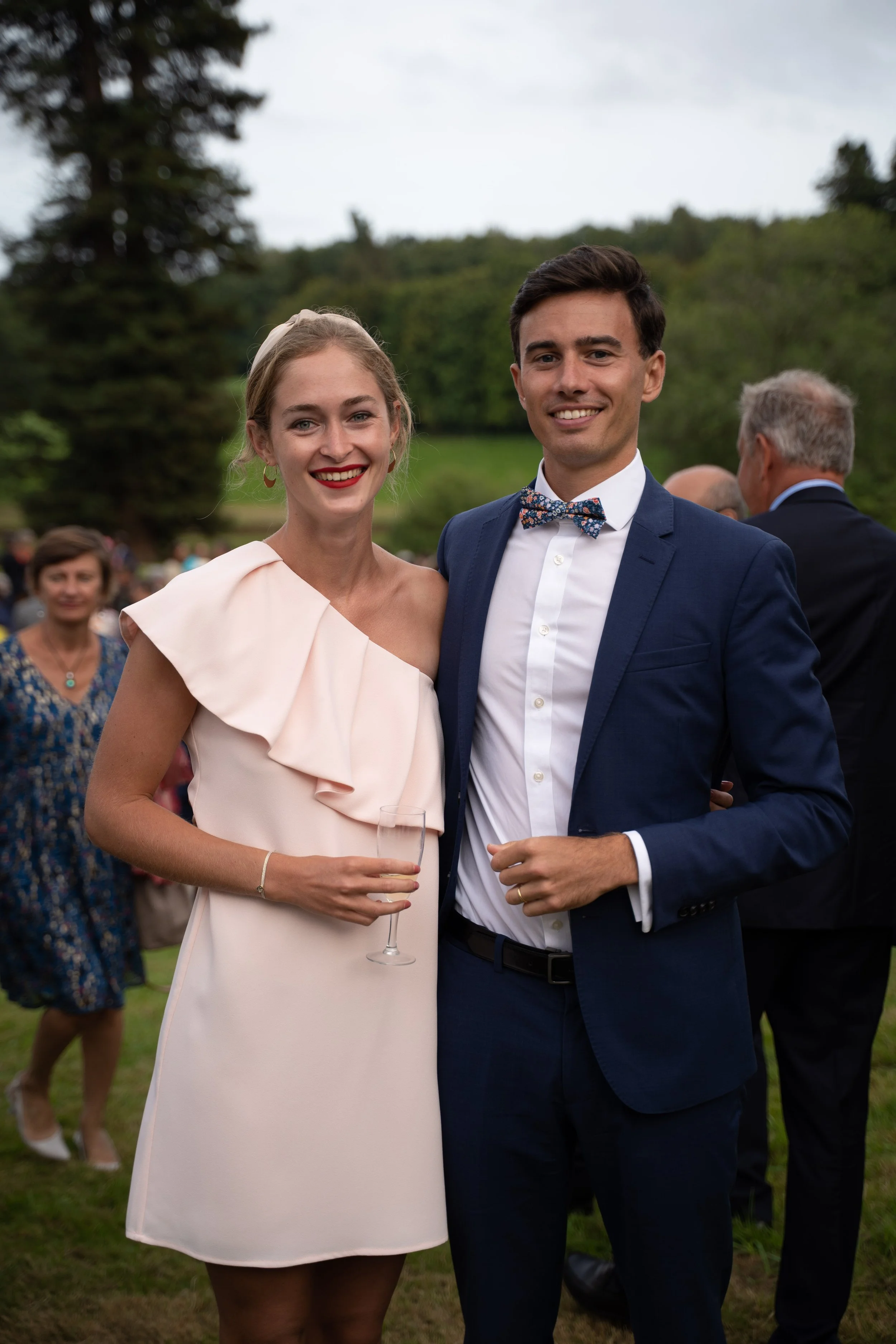 Un couple souriant lors d'une réception en plein air. La femme porte une robe pâle avec une épaule dénudée, tenant un verre à champagne. L'homme porte un costume bleu foncé avec un nœud papillon à motifs. En arrière-plan, d'autres personnes en vêteme