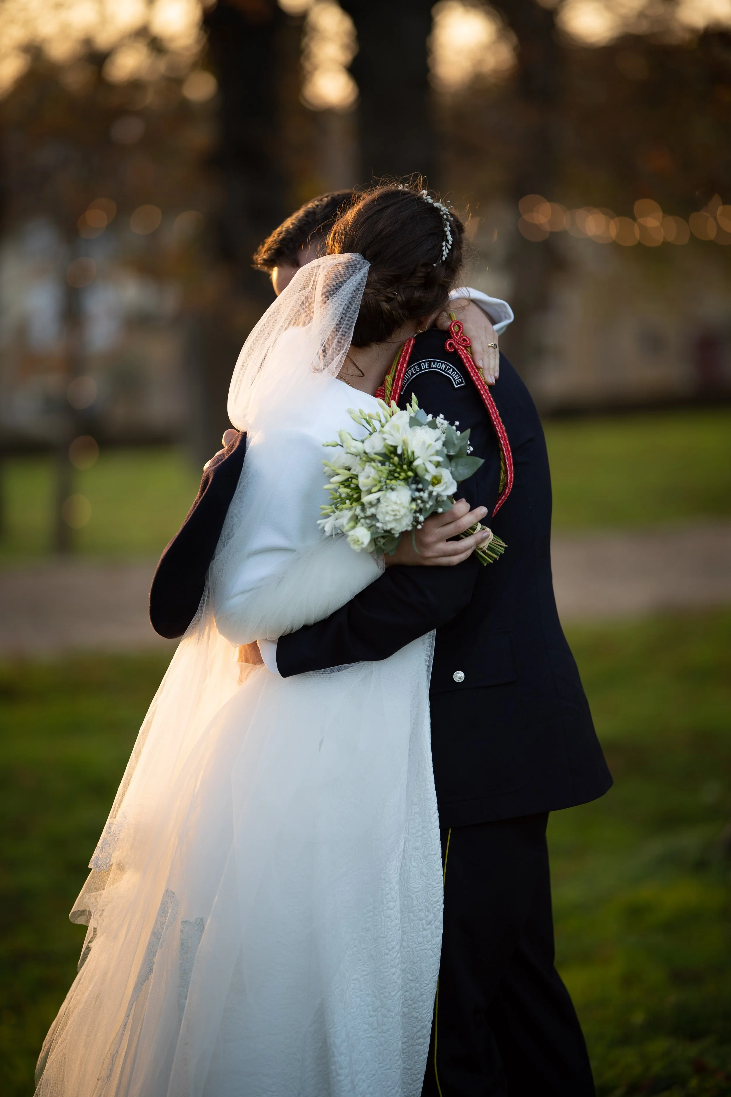 Un couple en habits de mariage s'embrassant dans un parc lors du coucher du soleil, la femme tenant un bouquet de fleurs blanches.