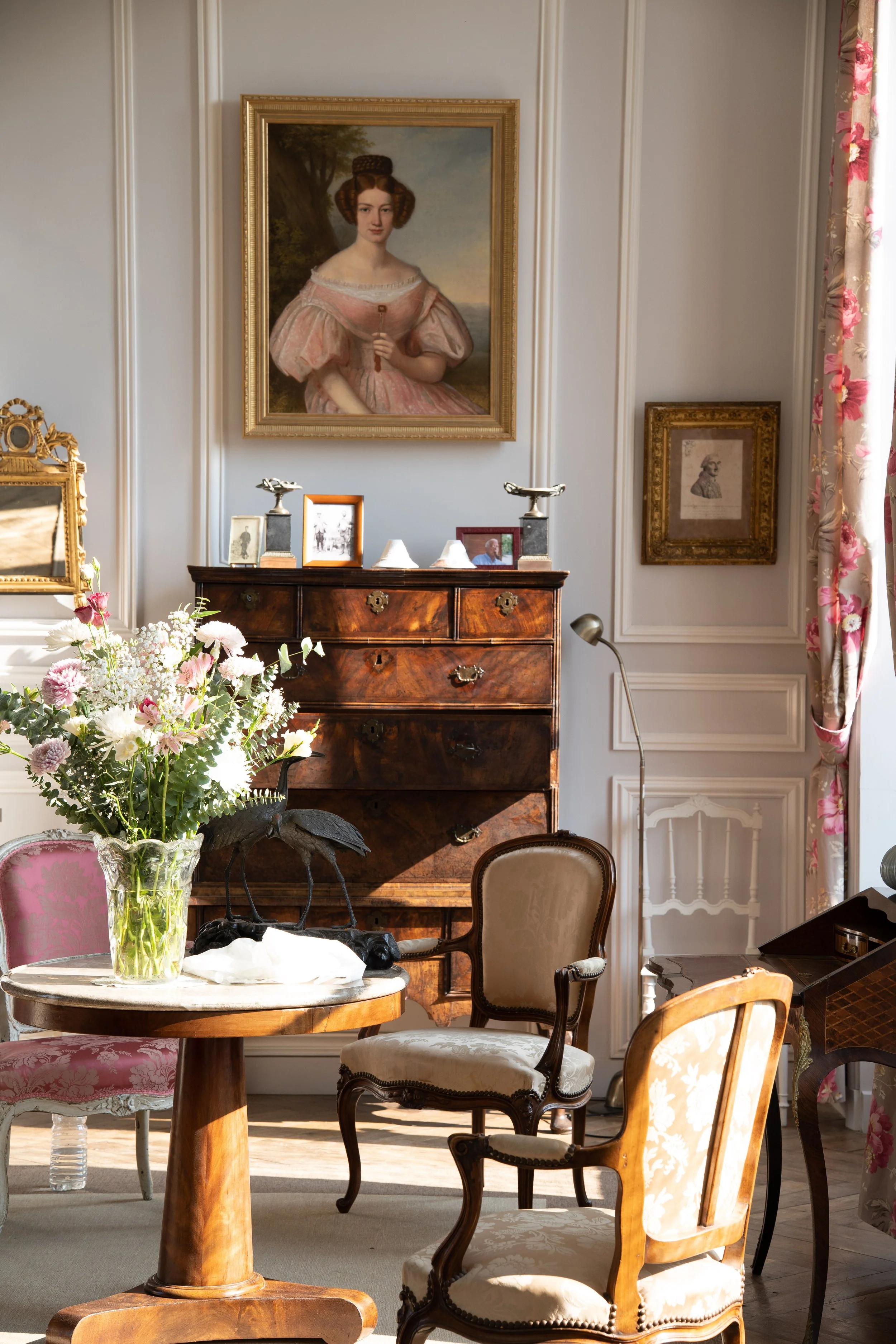 Salon élégant avec un tableau de femme dans un cadre doré, une commode en bois, des fleurs dans un vase, plusieurs chaises anciennes en bois et tissu, rideaux fleuris, et un lampadaire en métal.