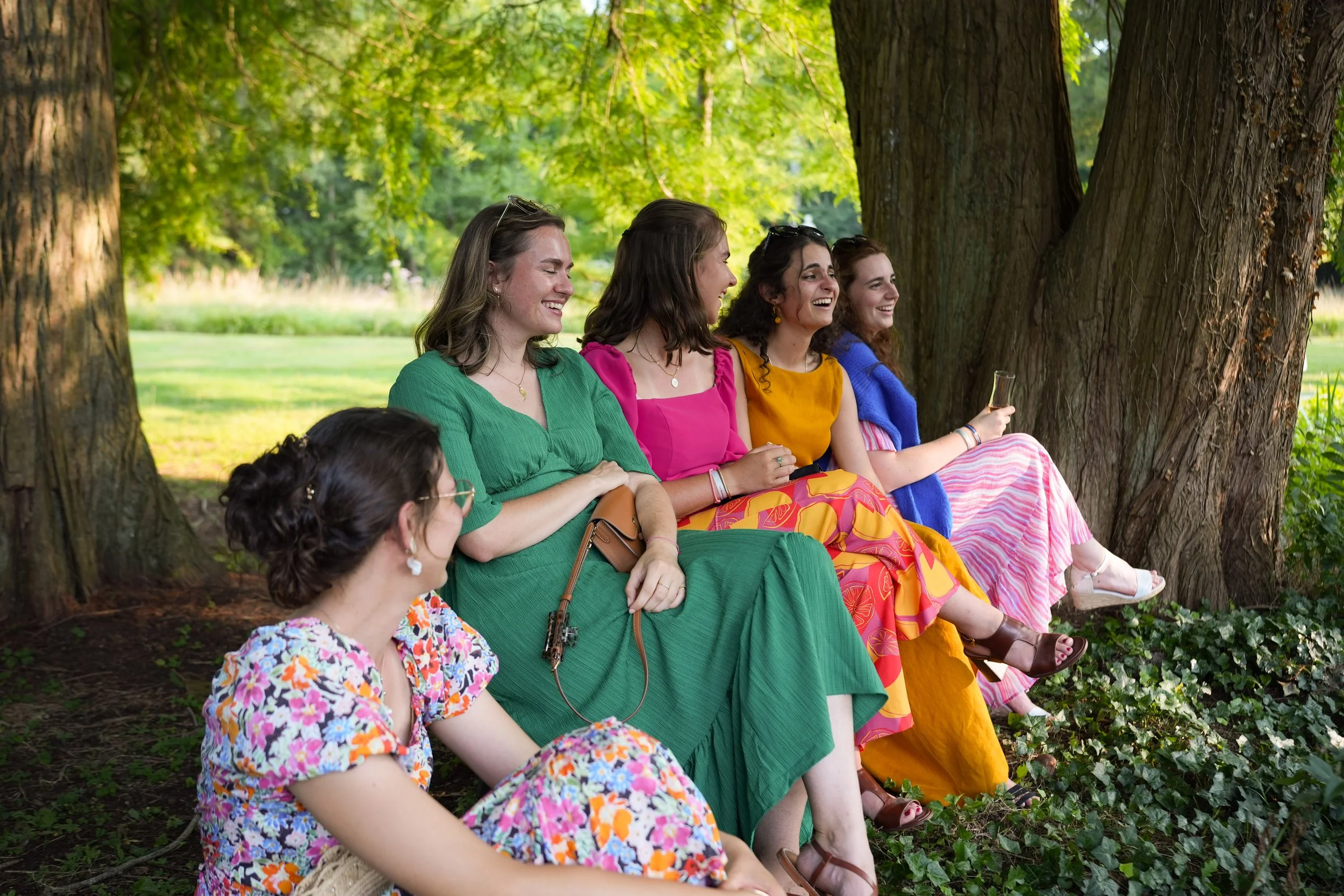 Groupe de femmes assises sous un arbre dans un parc, souriantes et discutant, avec des vêtements colorés.