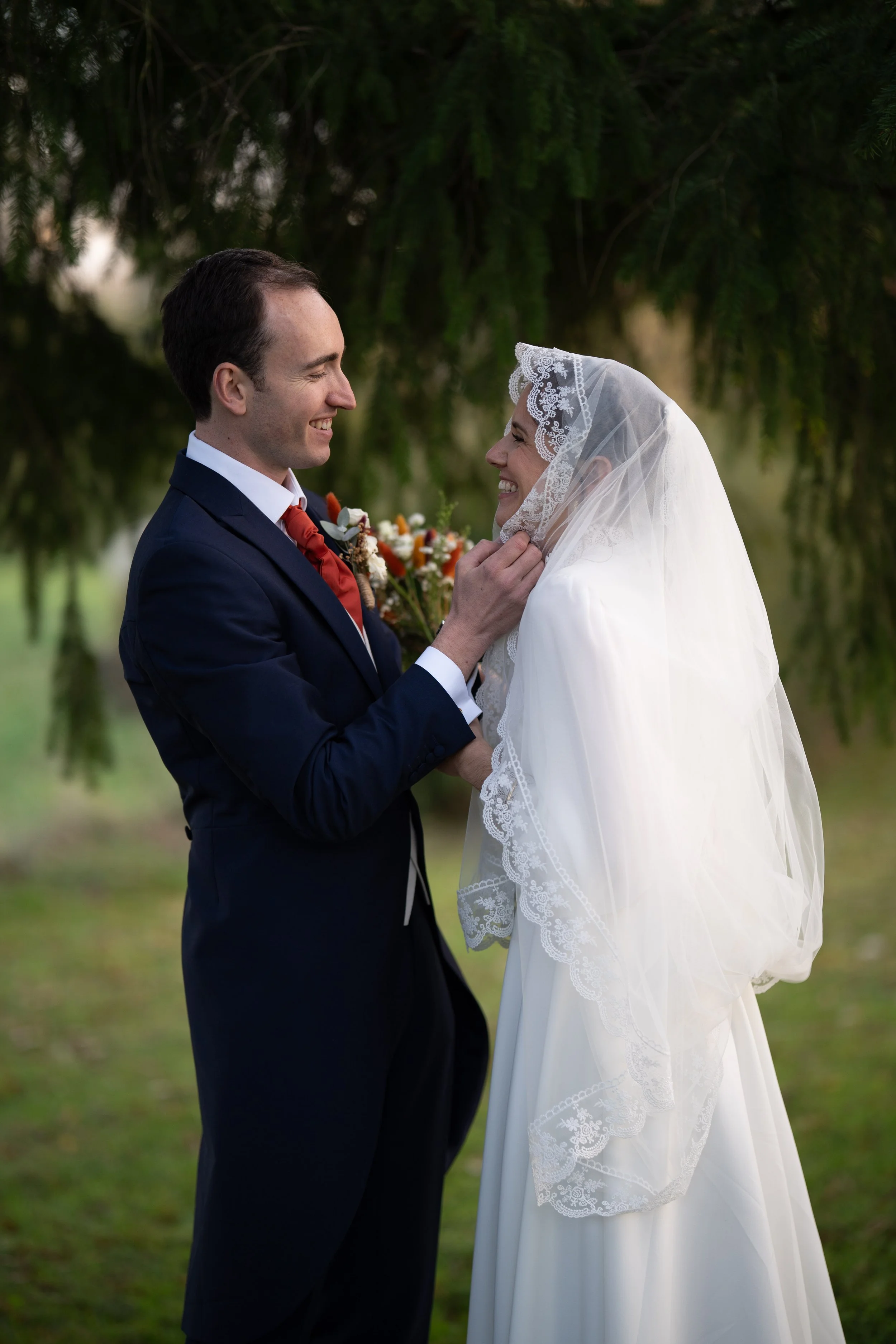 Un homme en costume souriant regarde une femme dans une robe de mariage, qui sourit également. Ils sont dans un environnement extérieur avec des arbres en arrière-plan.