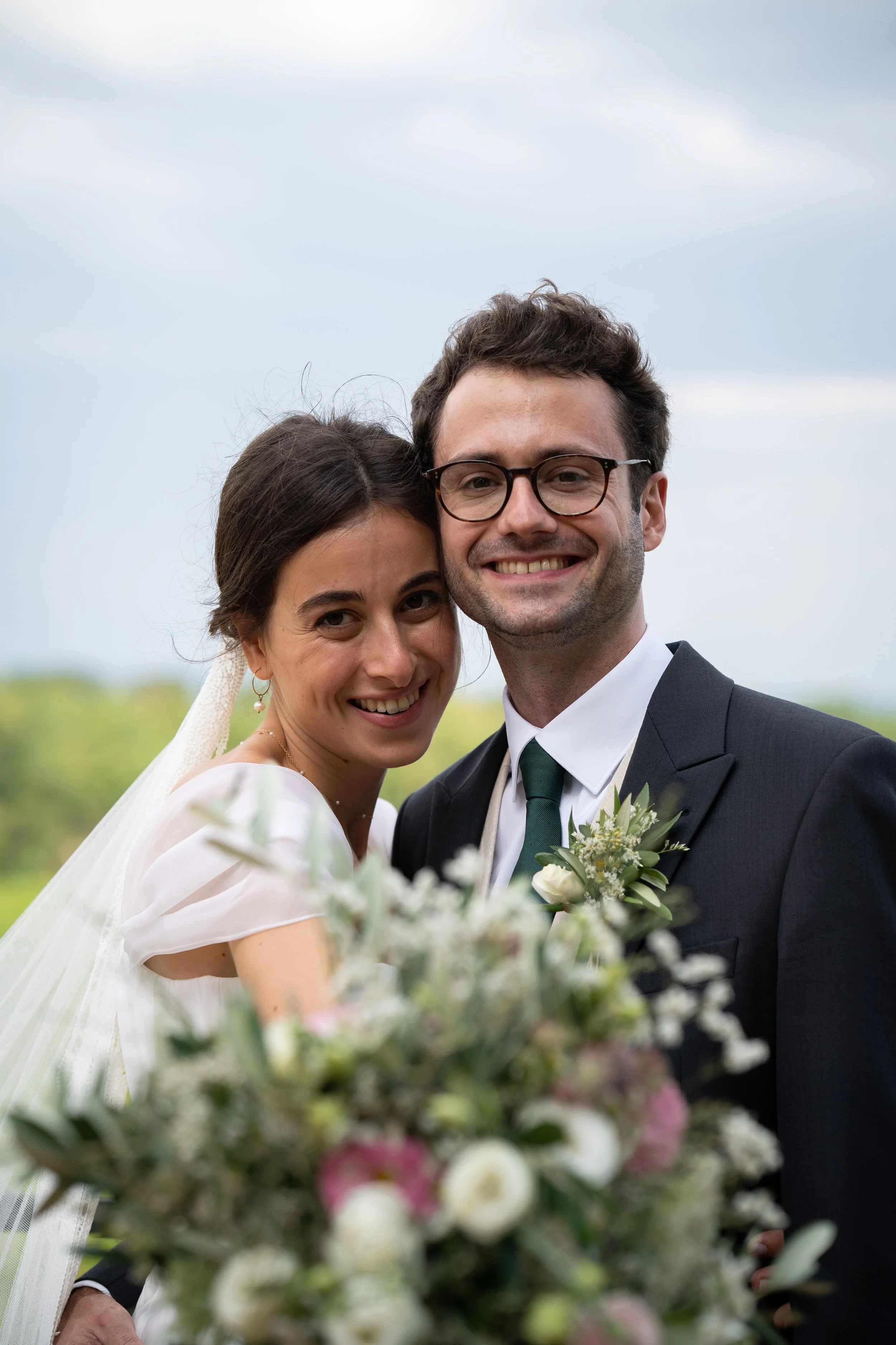 Un couple marié souriant, la femme en robe de mariage et le homme en costume, tenant un bouquet de fleurs, lors d'un mariage en plein air avec un ciel nuageux en arrière-plan.