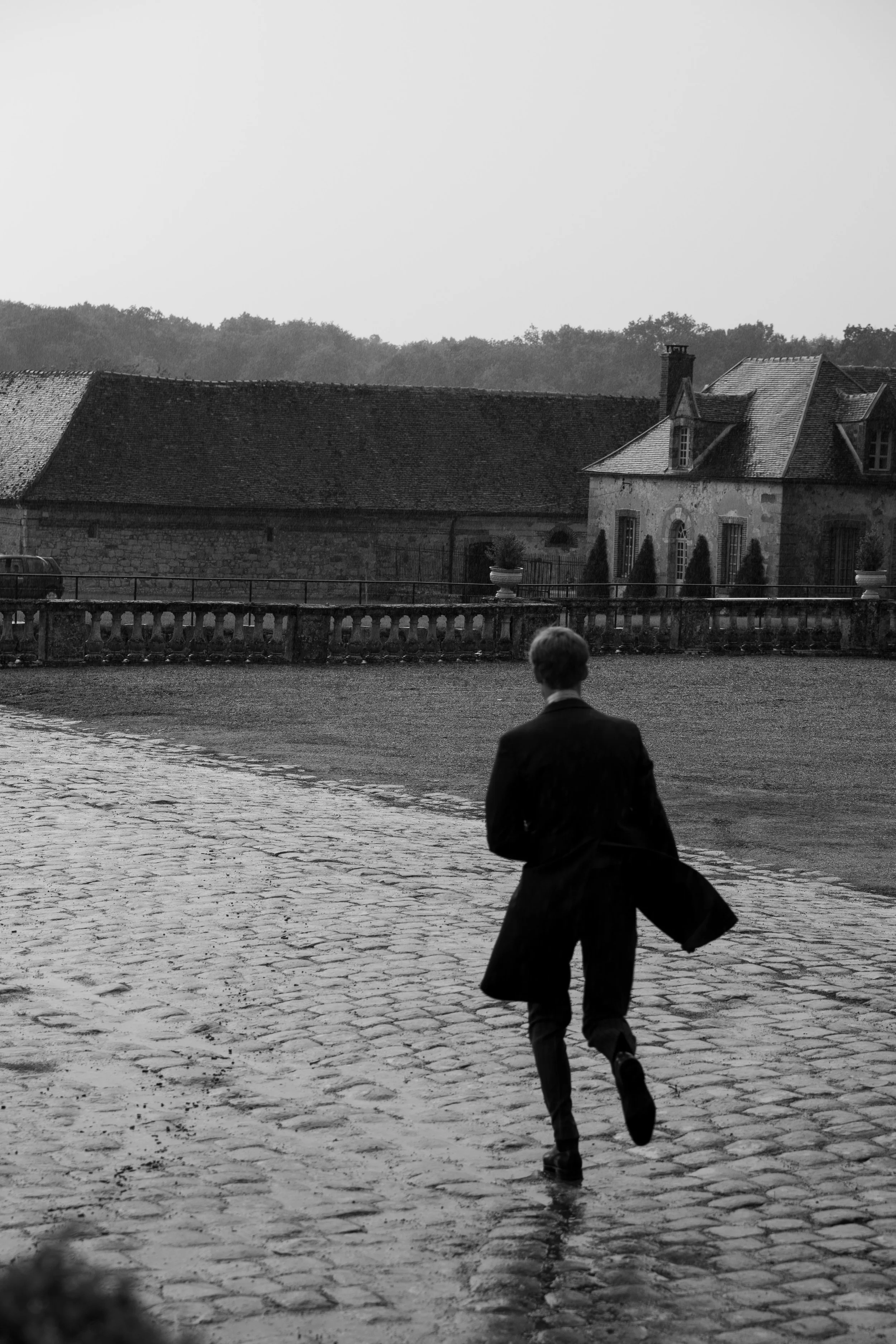 Une personne habillée en costume noir court sur un trottoir en pierre, avec un bâtiment ancien en pierre et une rampe en fer forgé en arrière-plan. La scène est en noir et blanc.