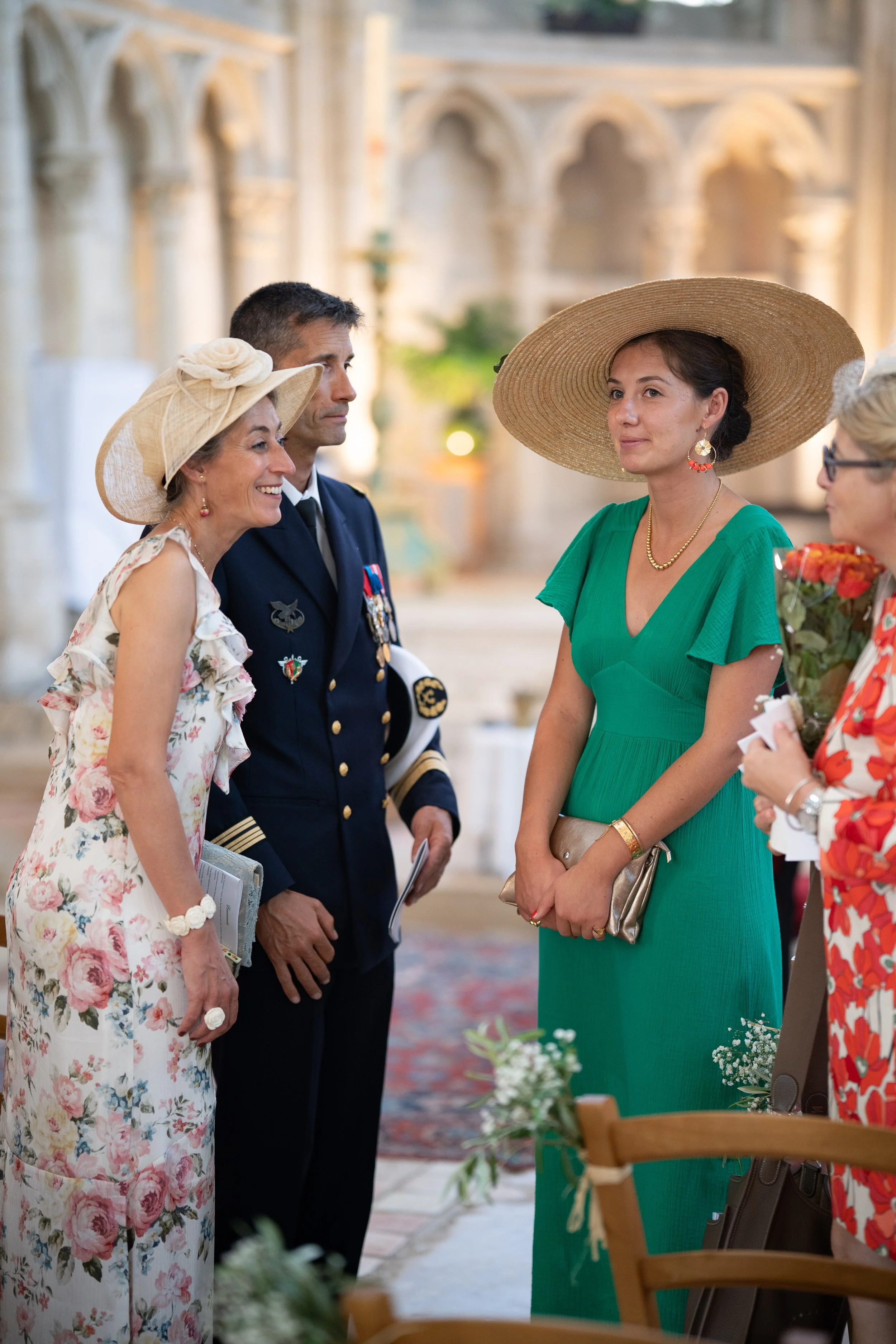 Cinq personnes lors d'une cérémonie, dont quatre femmes portant des habillements colorés et un homme en uniforme militaire. Deux femmes portent des chapeaux, et une tient un bouquet de fleurs. Ils se tiennent dans une salle ornée de sculptures et d'a