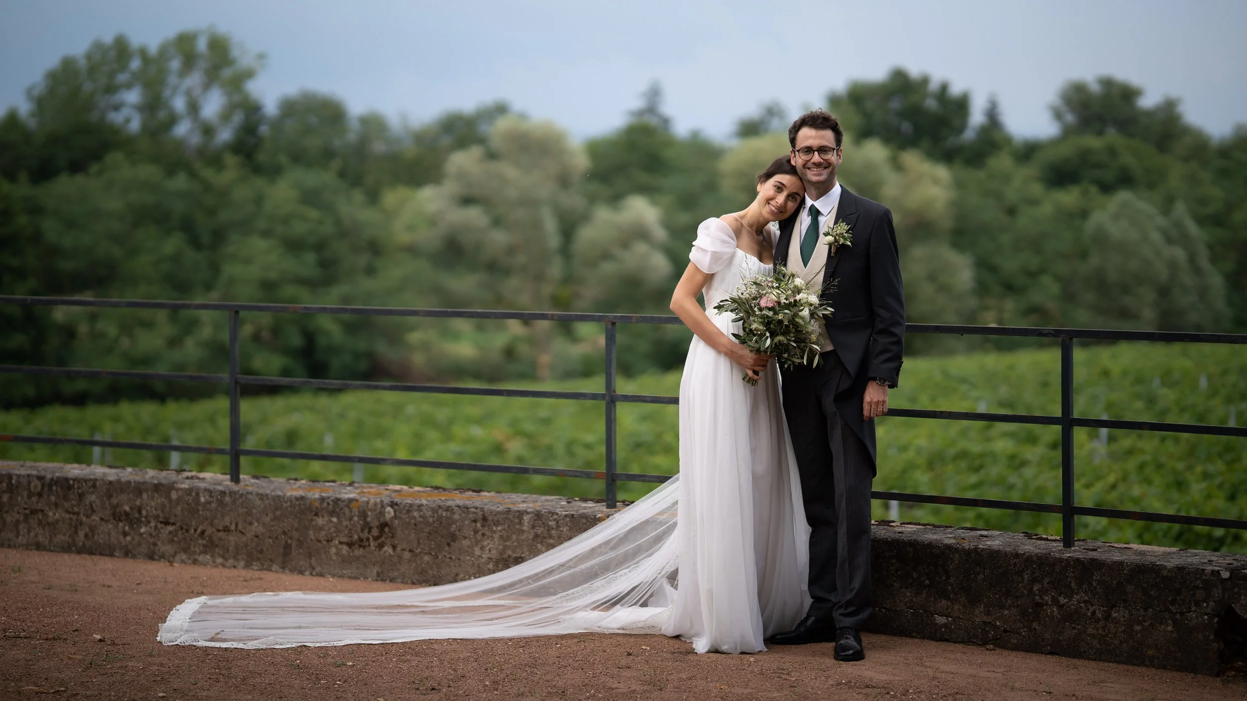 Un couple de mariés, un homme et une femme, sourient et se tiennent ensemble devant un paysage verdoyant. La femme porte une robe de mariée blanche avec un long train, et l'homme porte un costume noir avec une cravate verte. La femme tient un bouquet