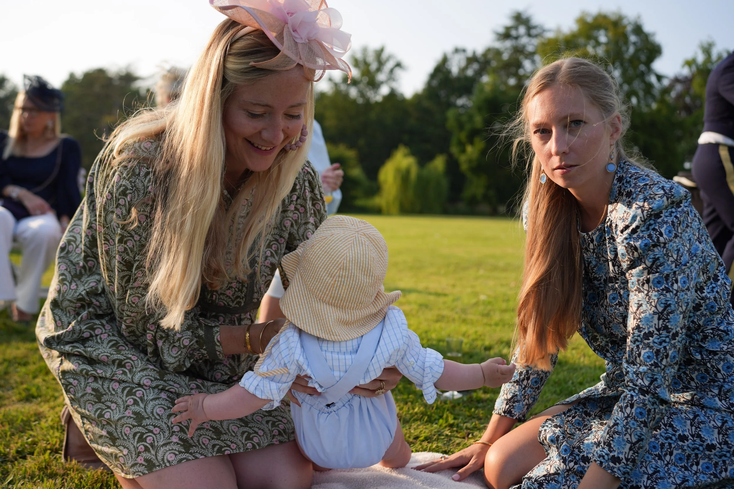 Un groupe de femmes et un enfant en train de jouer à l'extérieur lors d'une journée ensoleillée dans un parc. Deux femmes portent des vêtements à motif floral et un chapeau orné de rubans, tandis que l'enfant, habillé en vêtements légers avec une che